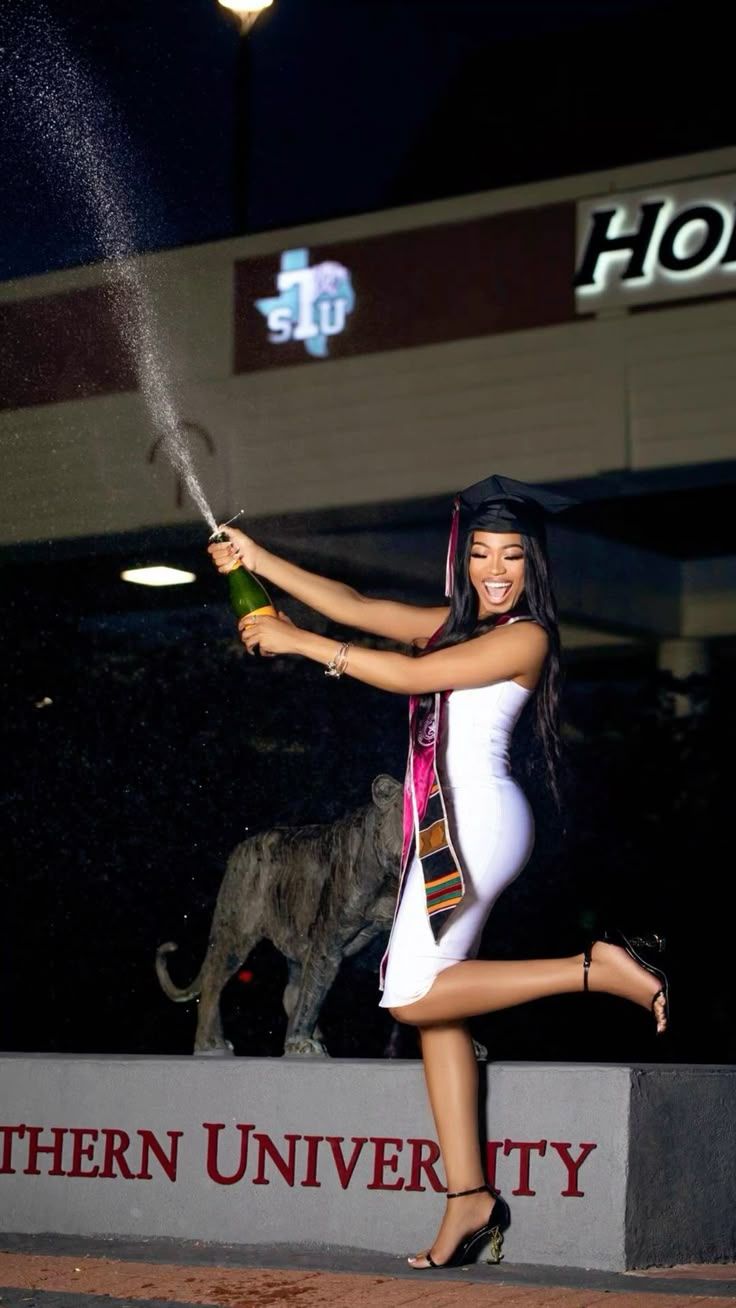 Graduate celebrates near university sign at night with champagne spray while wearing white dress and graduation cap.