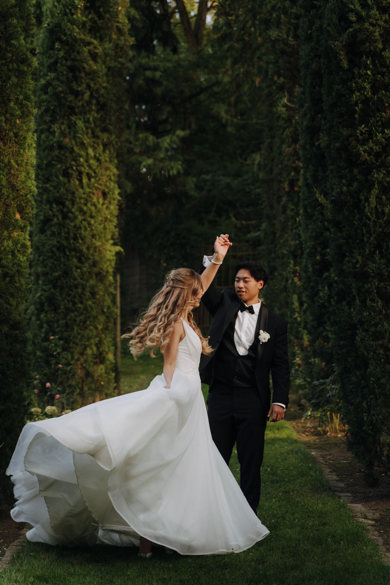 A romantic couple dancing and twirling in an elegant garden setting with tall trees and greenery at dusk.