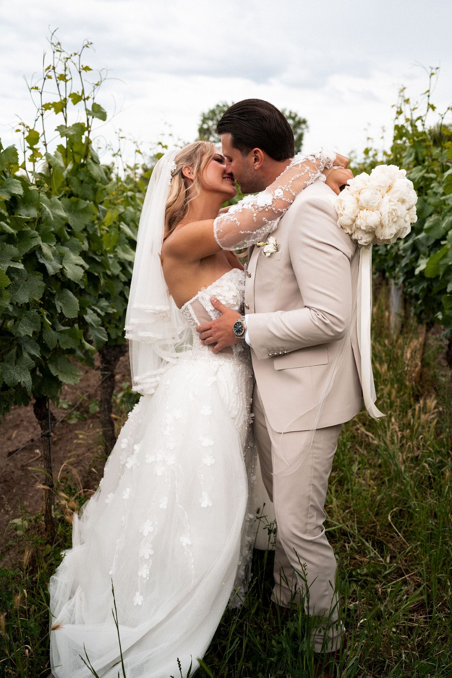 Bride and groom kissing in a vineyard, bride in white star-embroidered gown with veil, groom in beige suit holding bouquet.