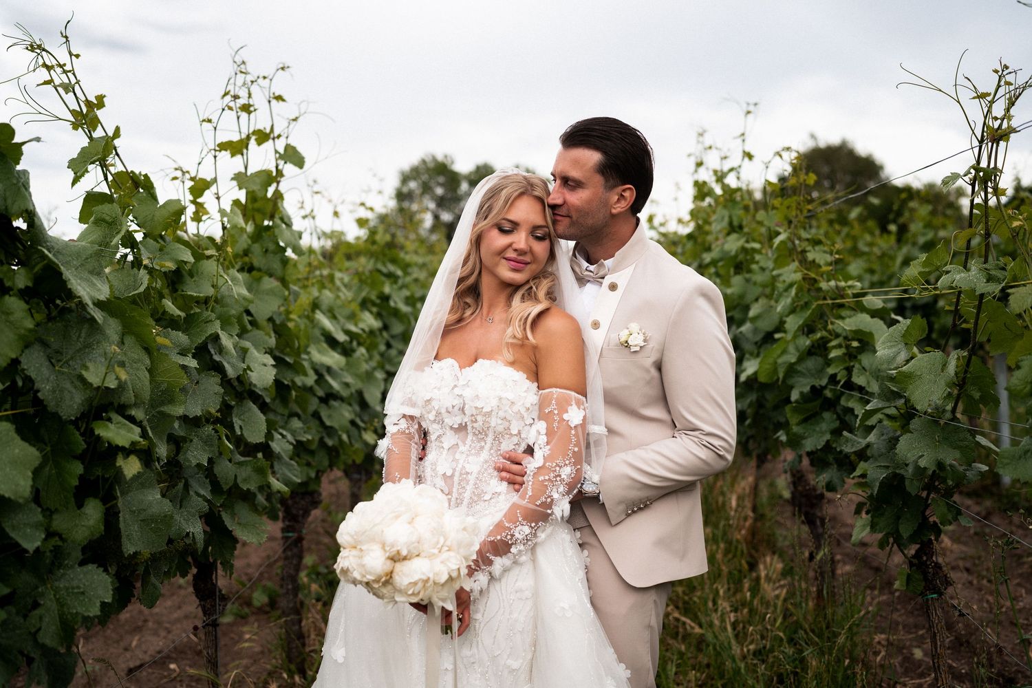 Bride and groom pose romantically among lush vineyard rows, she holds white peony bouquet in elegant lace wedding gown.