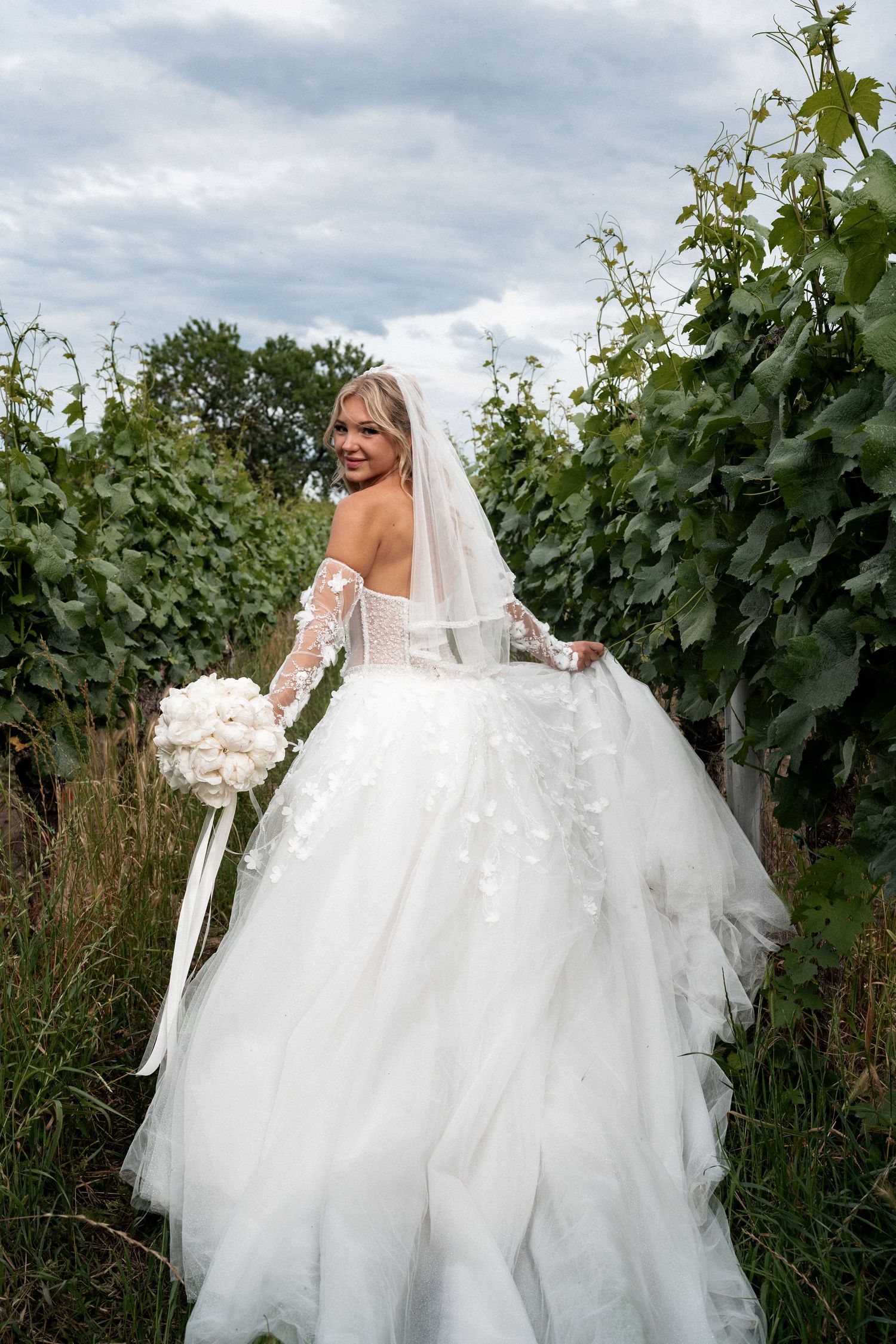 Bride in white lace ball gown with veil holds bouquet among vineyard rows, glancing back over shoulder.