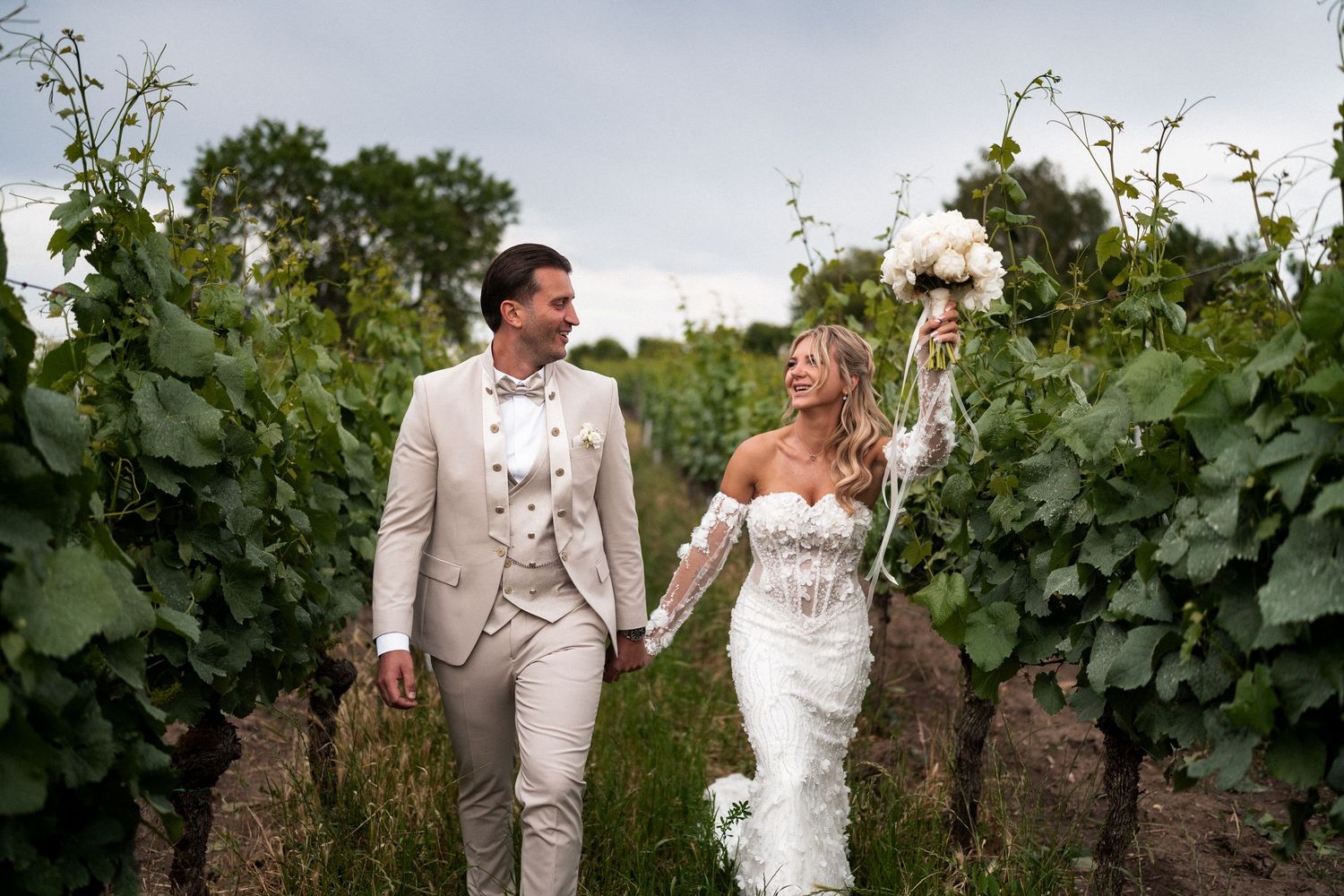 Bride and groom walk hand-in-hand through lush vineyard rows, she holds white bouquet aloft in elegant lace gown.