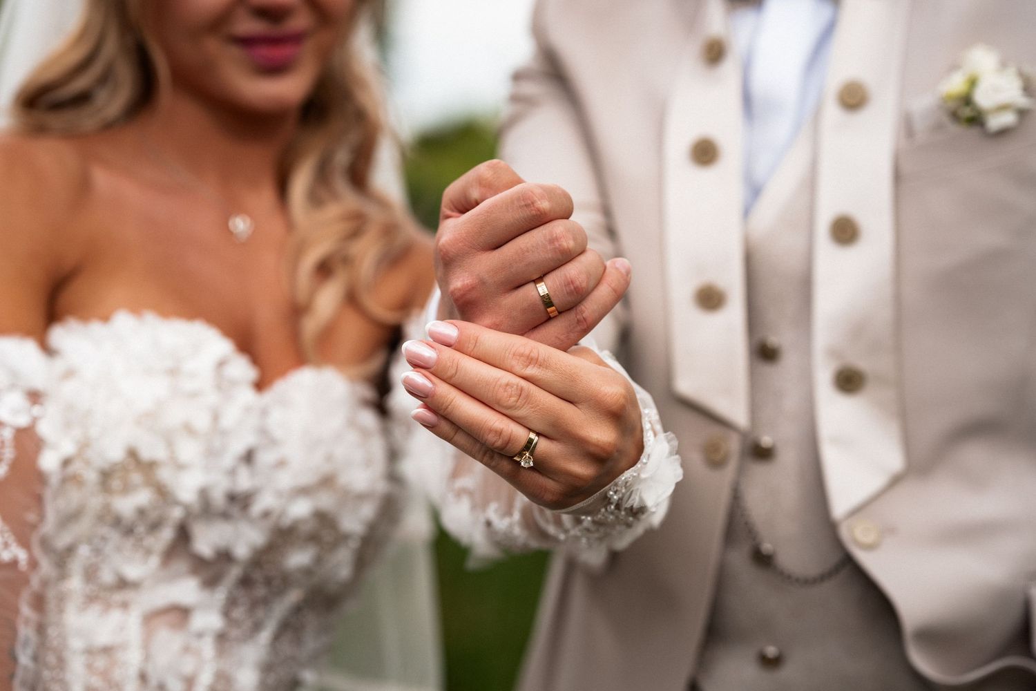 Bride and groom showing off wedding rings, bride in lace gown, groom in beige suit with boutonniere.