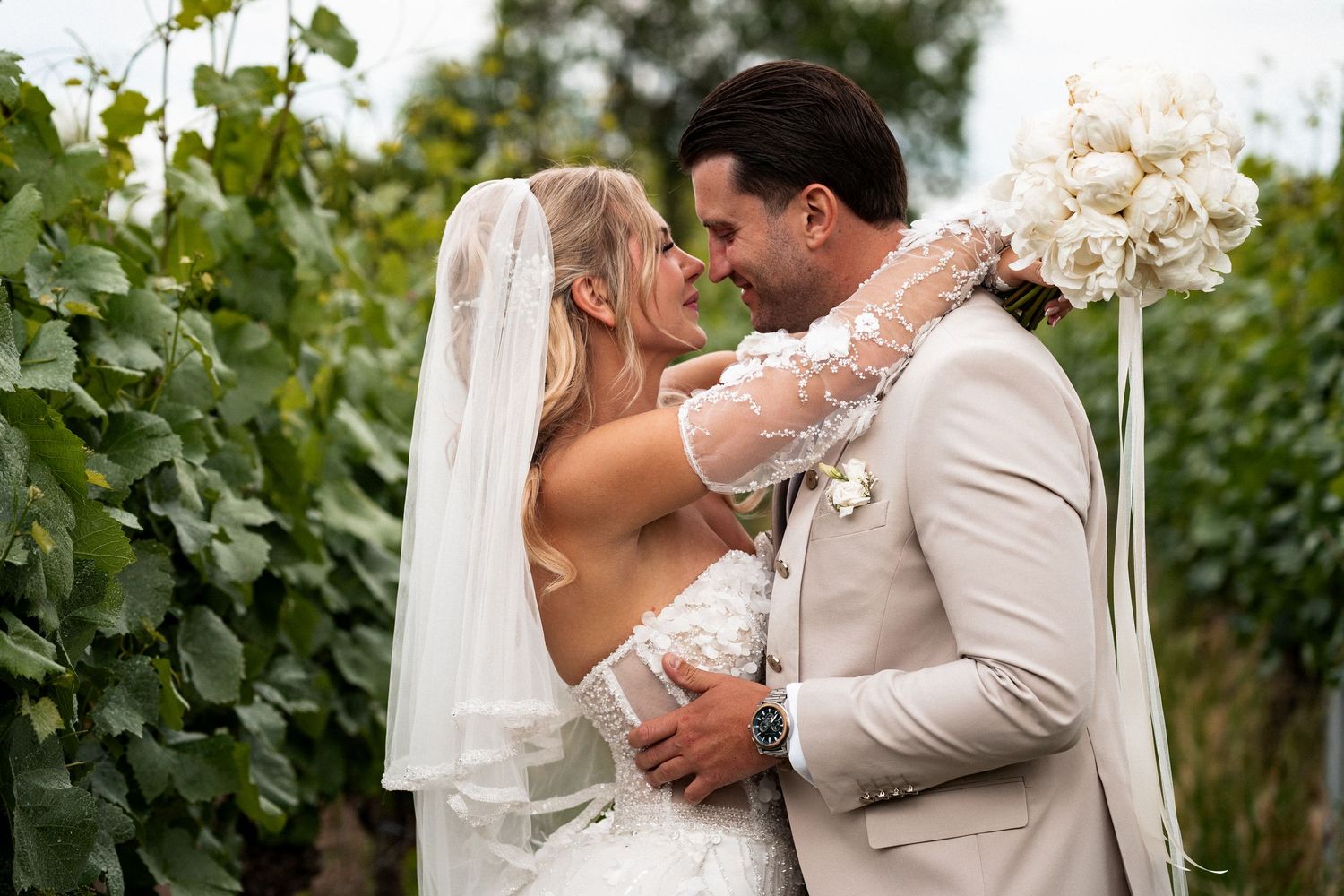 Bride and groom share romantic moment in vineyard, bride in lace veil holding white bouquet, groom in beige suit.