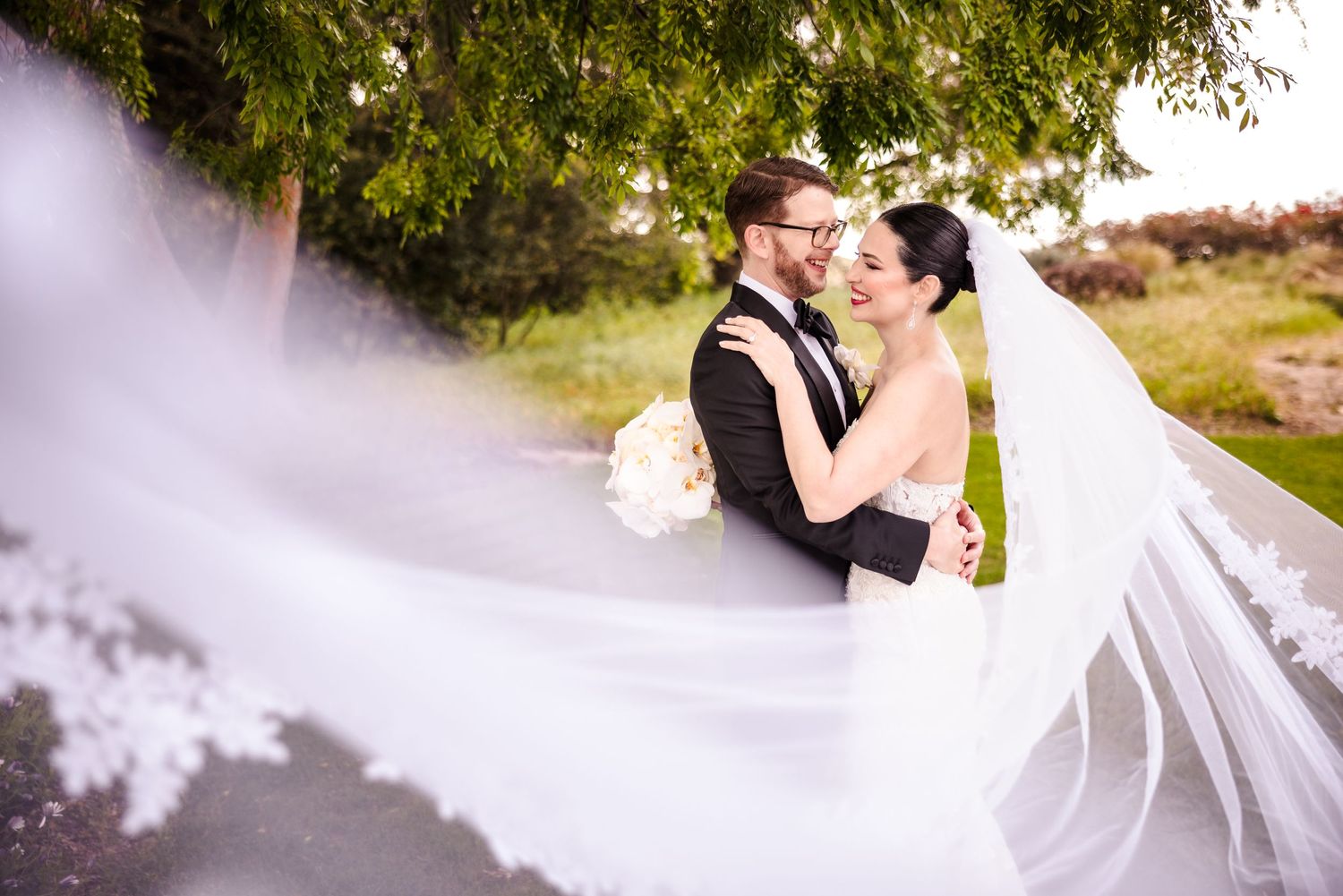 A romantic wedding couple embraces on a pathway as the bride's veil flows dramatically in the breeze.