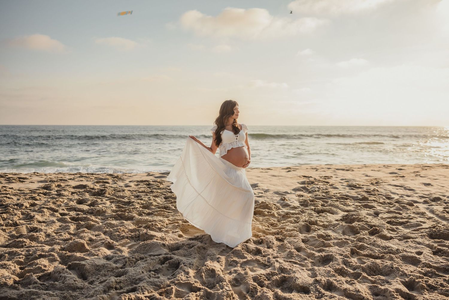 A person in a flowing white dress stands on a sandy beach at sunset while waves roll in from the ocean.
