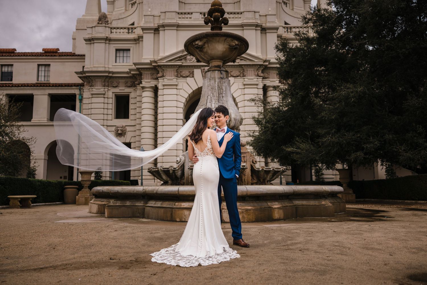 A couple in wedding attire embraces in front of an ornate fountain at a grand historical building.