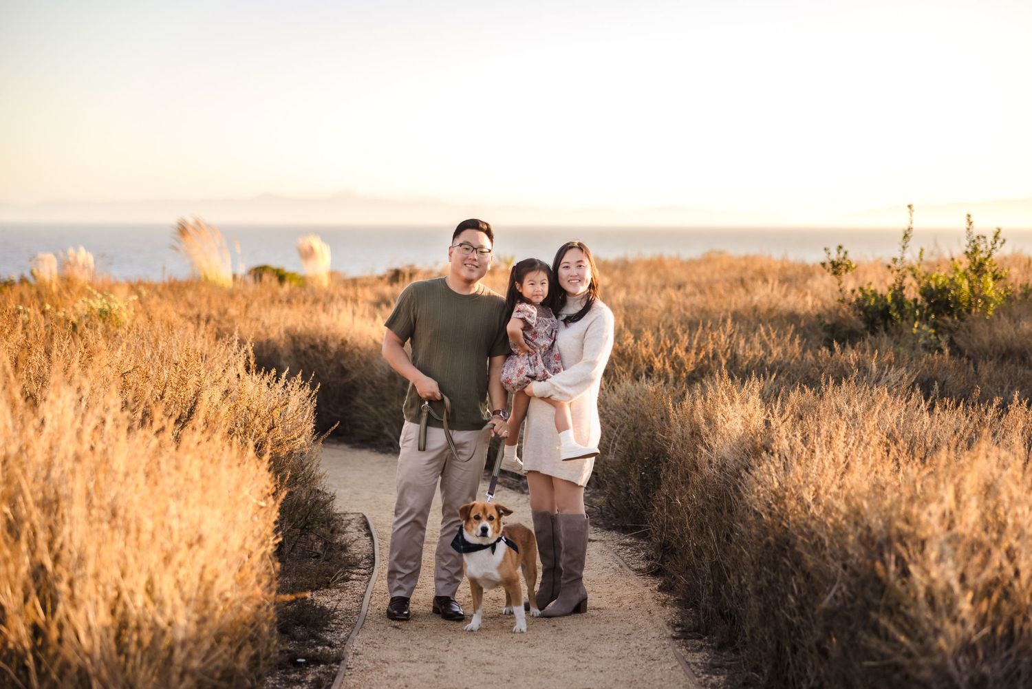 A family and their dog walk together on a trail through golden grass at sunset.