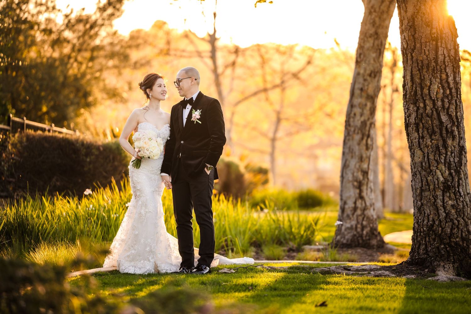 A couple in wedding attire stands together in a park at golden hour with sunlight streaming through trees.
