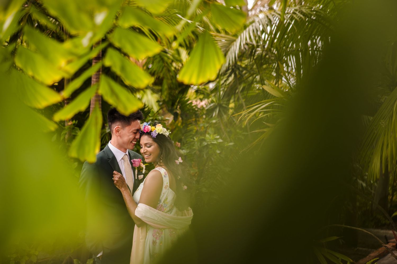 A romantic wedding couple embraces in a lush green garden setting framed by tropical foliage.