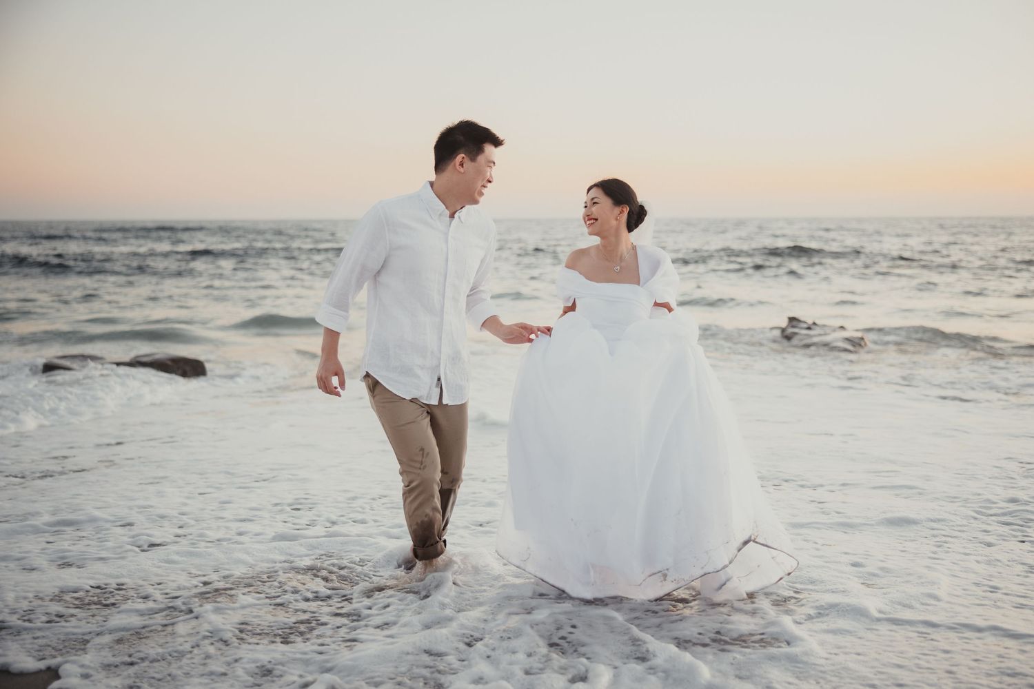 A couple walks hand in hand on a sandy beach at sunset wearing white attire and wedding dress.