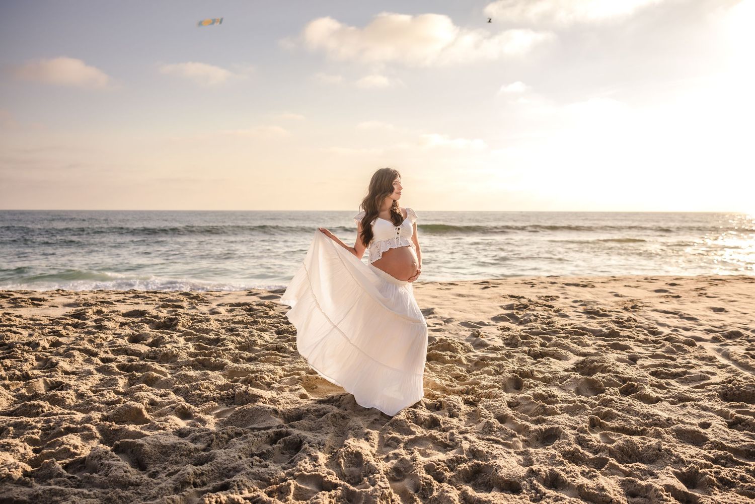 A person in a white dress stands on a beach at sunset with waves creating a serene coastal backdrop.