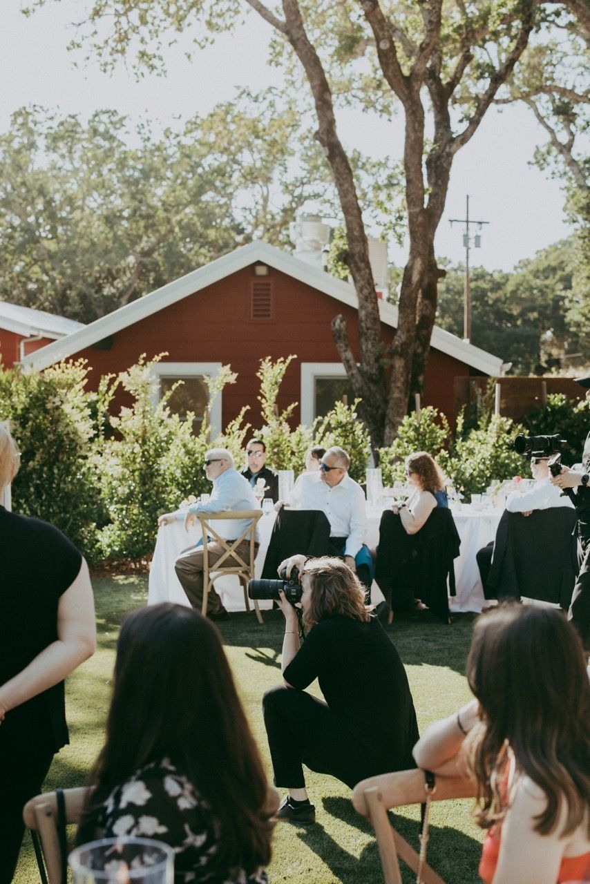 Outdoor wedding ceremony on green lawn with guests seated, photographer crouching, and red barn building in background.