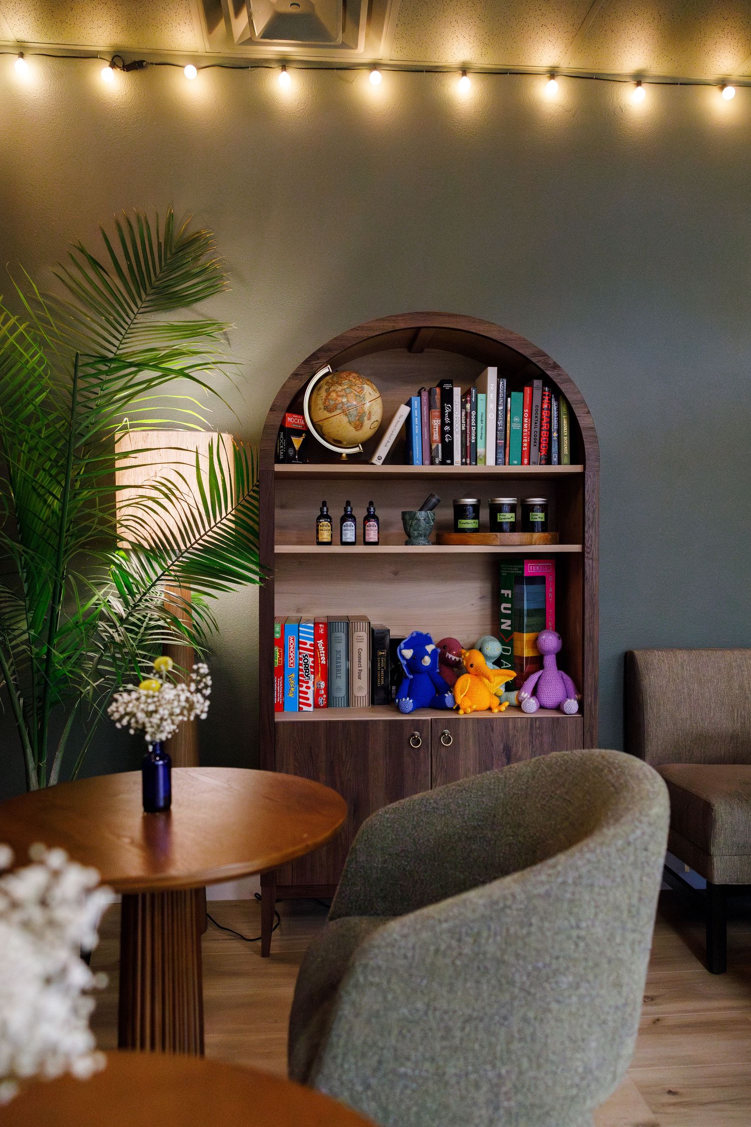 Cozy reading nook with arched bookshelf, string lights, palm plant and grey upholstered chairs around a wooden table.