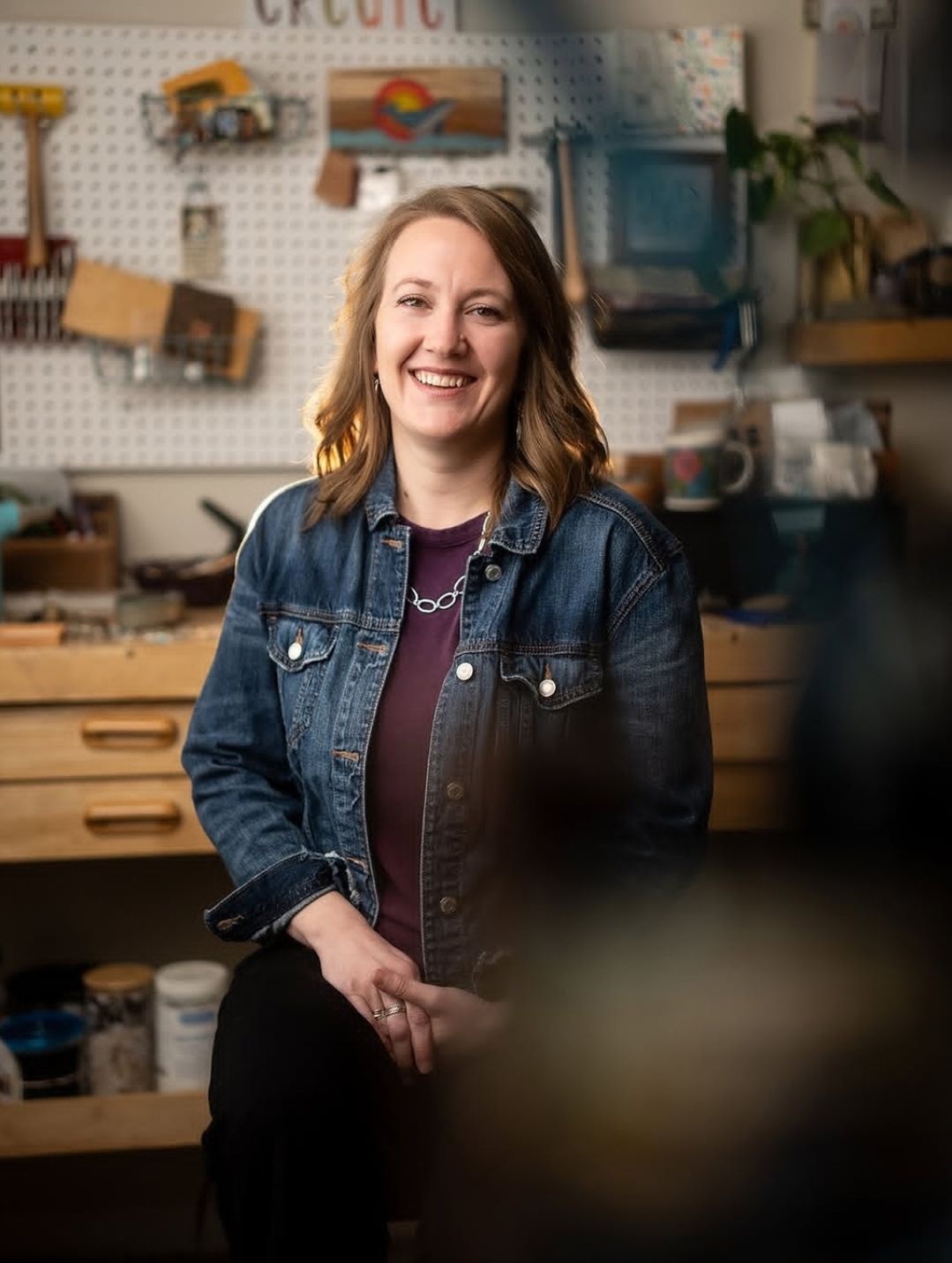 Smiling woman in denim jacket poses in a creative workshop with tools and pegboard wall behind her.