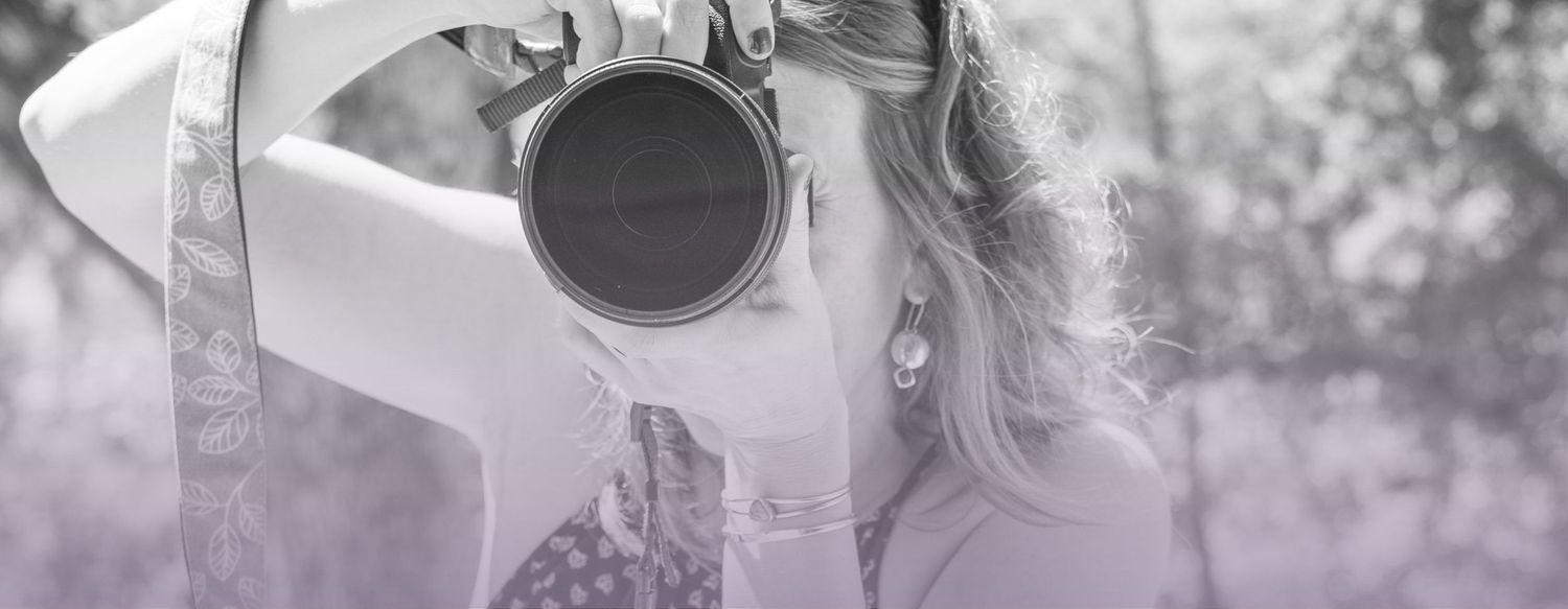 Black and white photo of a young woman holding a camera up to her face, photographing outdoors.