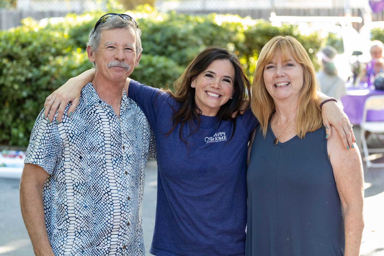 Three people stand together outdoors wearing casual attire and smiling on a sunny day.