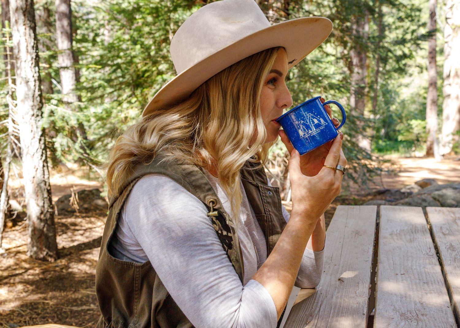 Woman in wide-brim hat sipping from blue campfire mug outdoors in a forest setting.