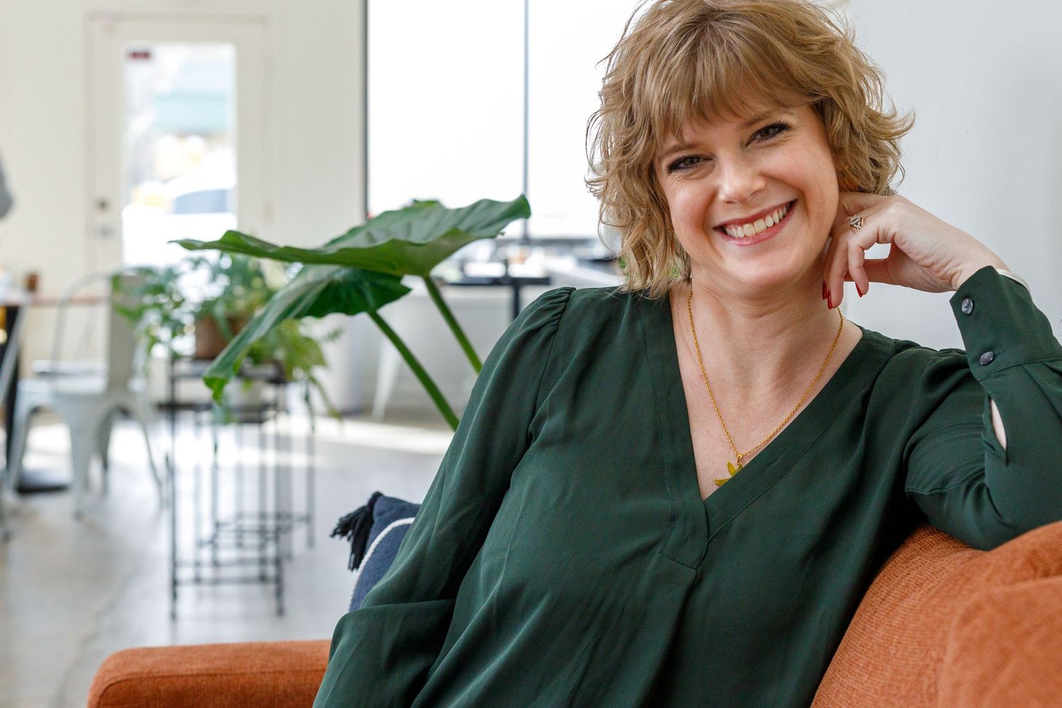 Relaxed figure in a green sweater sits on an orange couch while leaning against their hand in a bright interior space.
