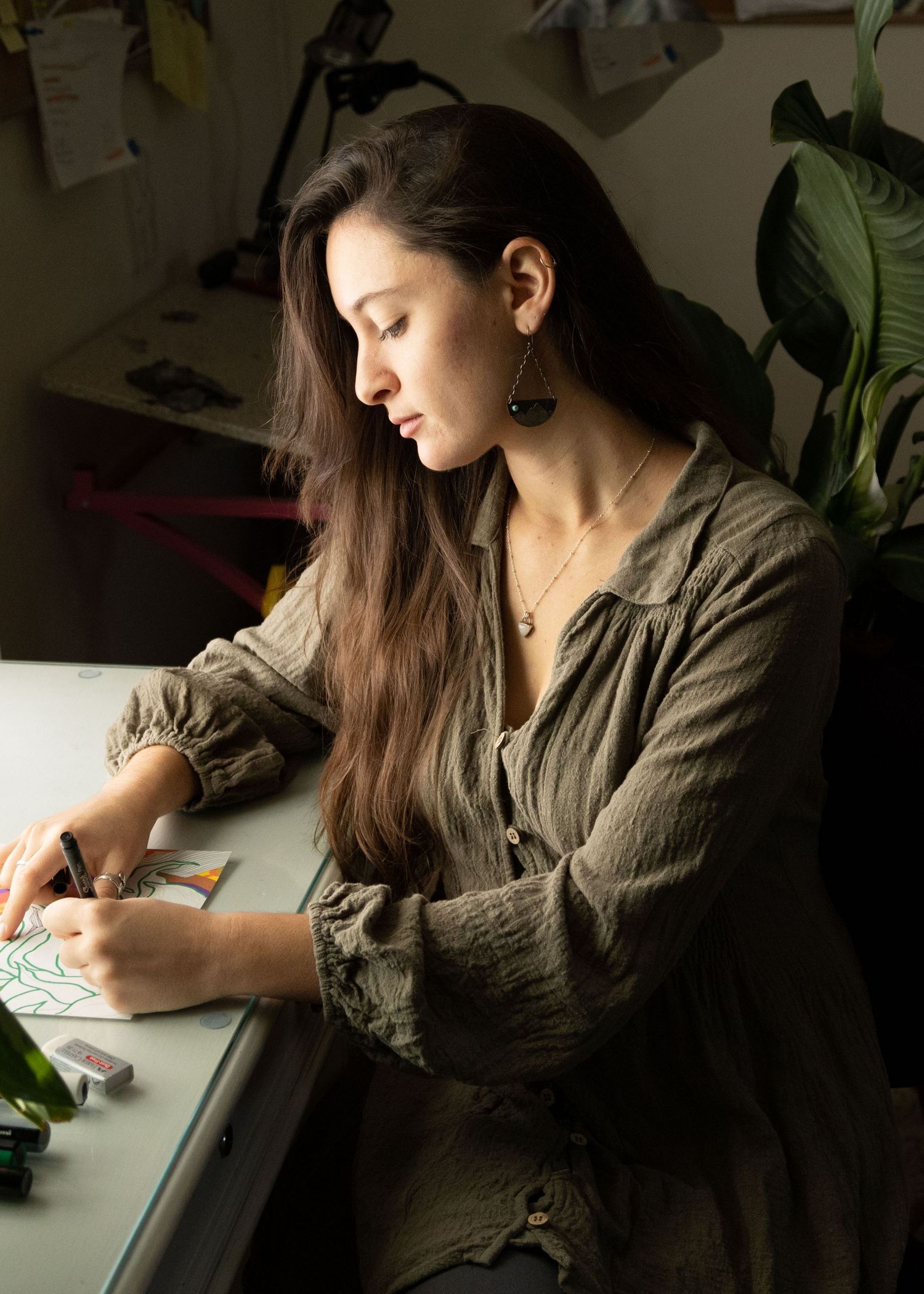 Person with long dark hair in an olive blouse sits at a desk writing near houseplants in natural lighting.