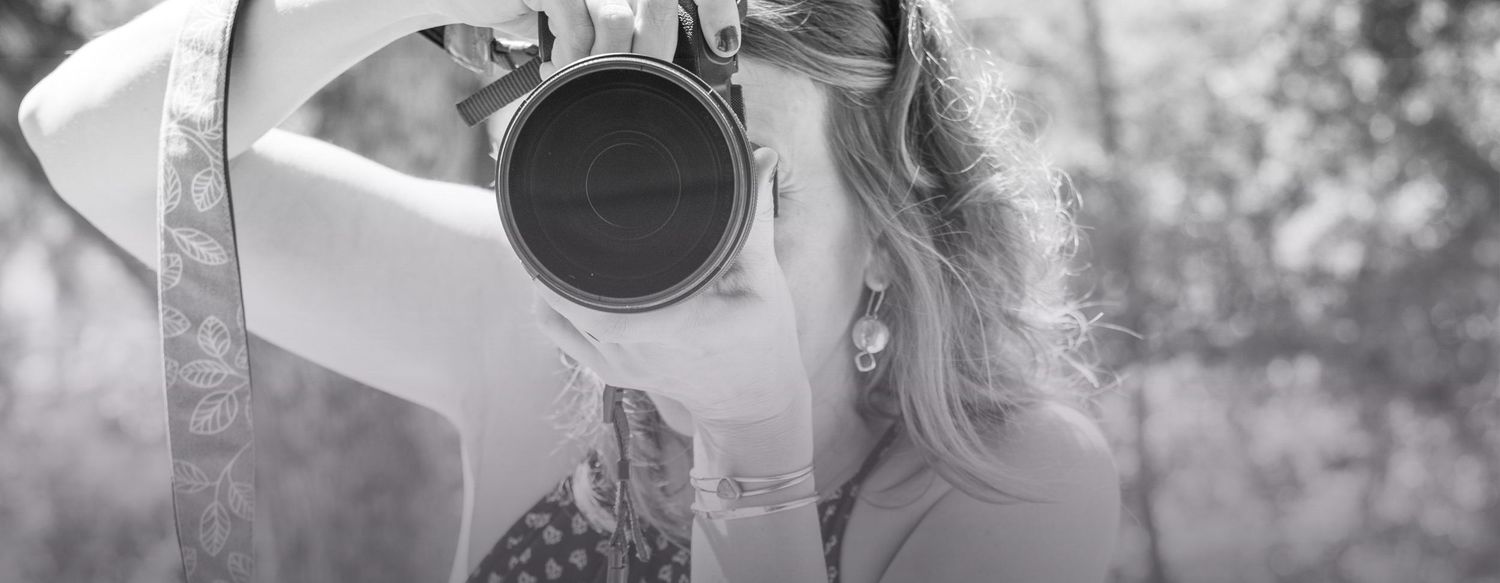 Black and white photo of a young woman with earrings holding a camera up to her face outdoors.