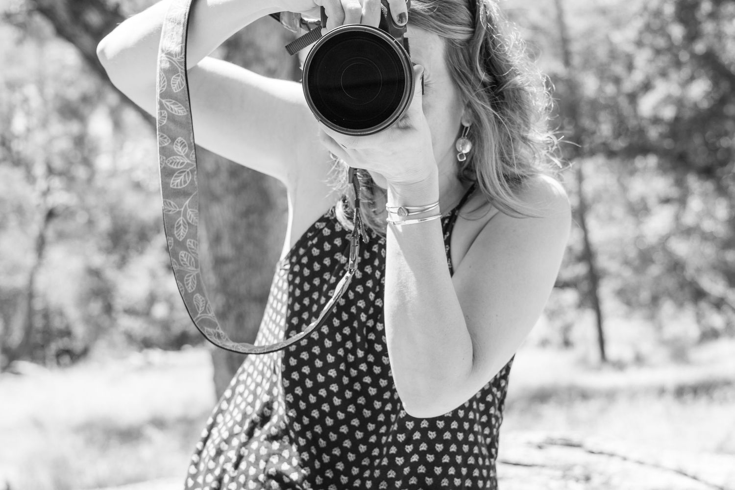Black and white photo of a woman in a polka dot dress photographing with a DSLR camera outdoors.