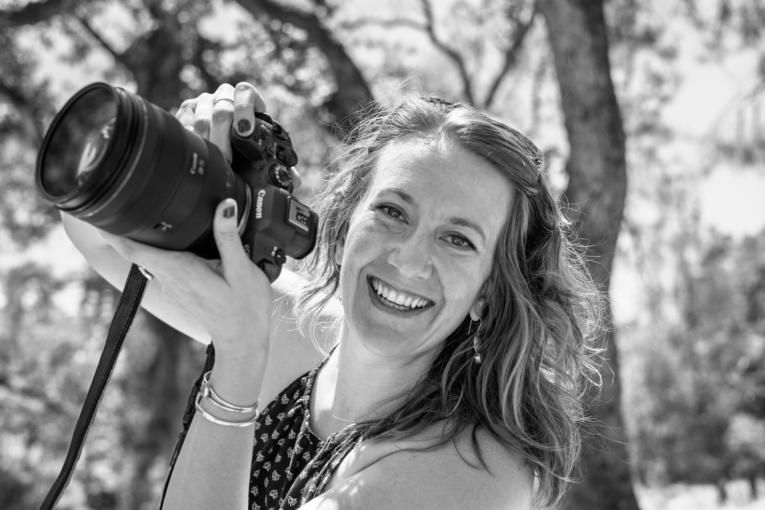 Woman smiling and holding a Canon camera outdoors in a black and white portrait photo.