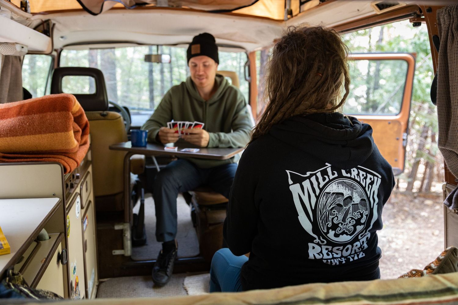 Two people sitting inside a vintage van with wood paneling having a conversation over coffee.
