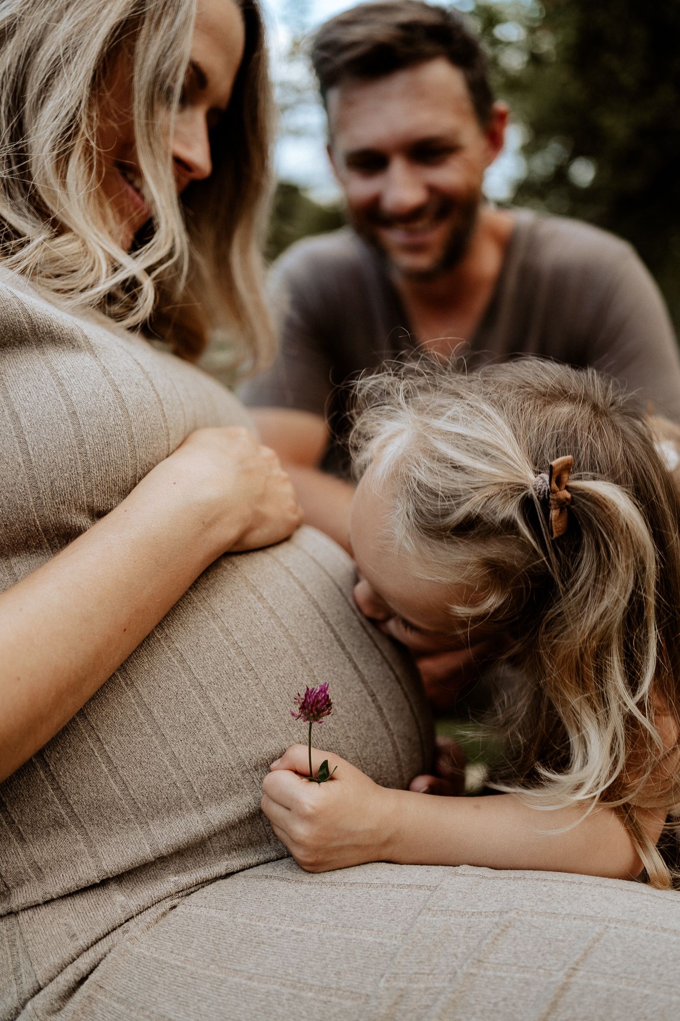 Natürliches Babybauch Shooting mit Familie im Freien, Kind küsst liebevoll den Bauch der Mutter.
