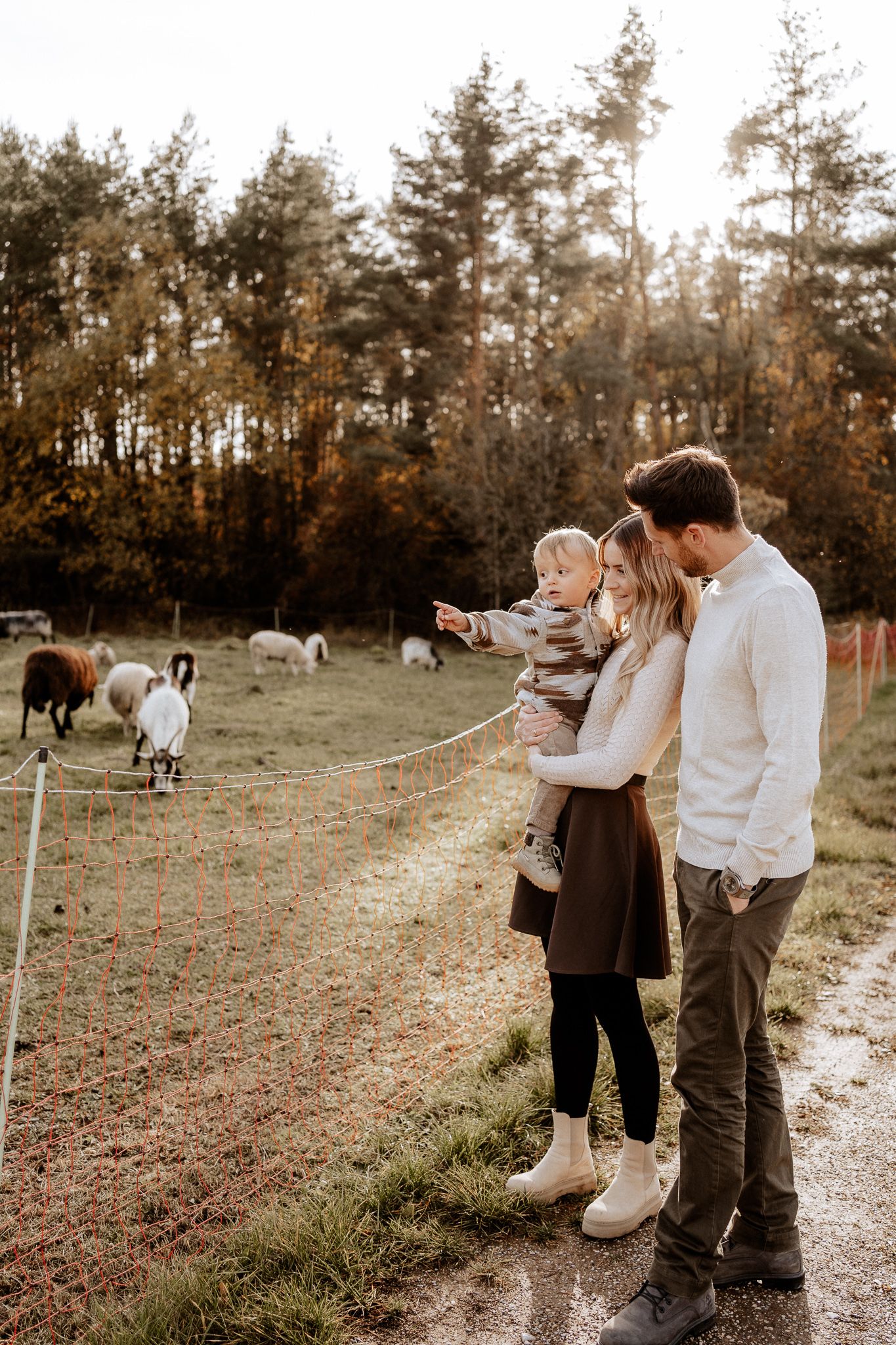 Familie steht gemeinsam am Zaun, während ihr Kind neugierig auf die Tiere auf der Wiese zeigt.