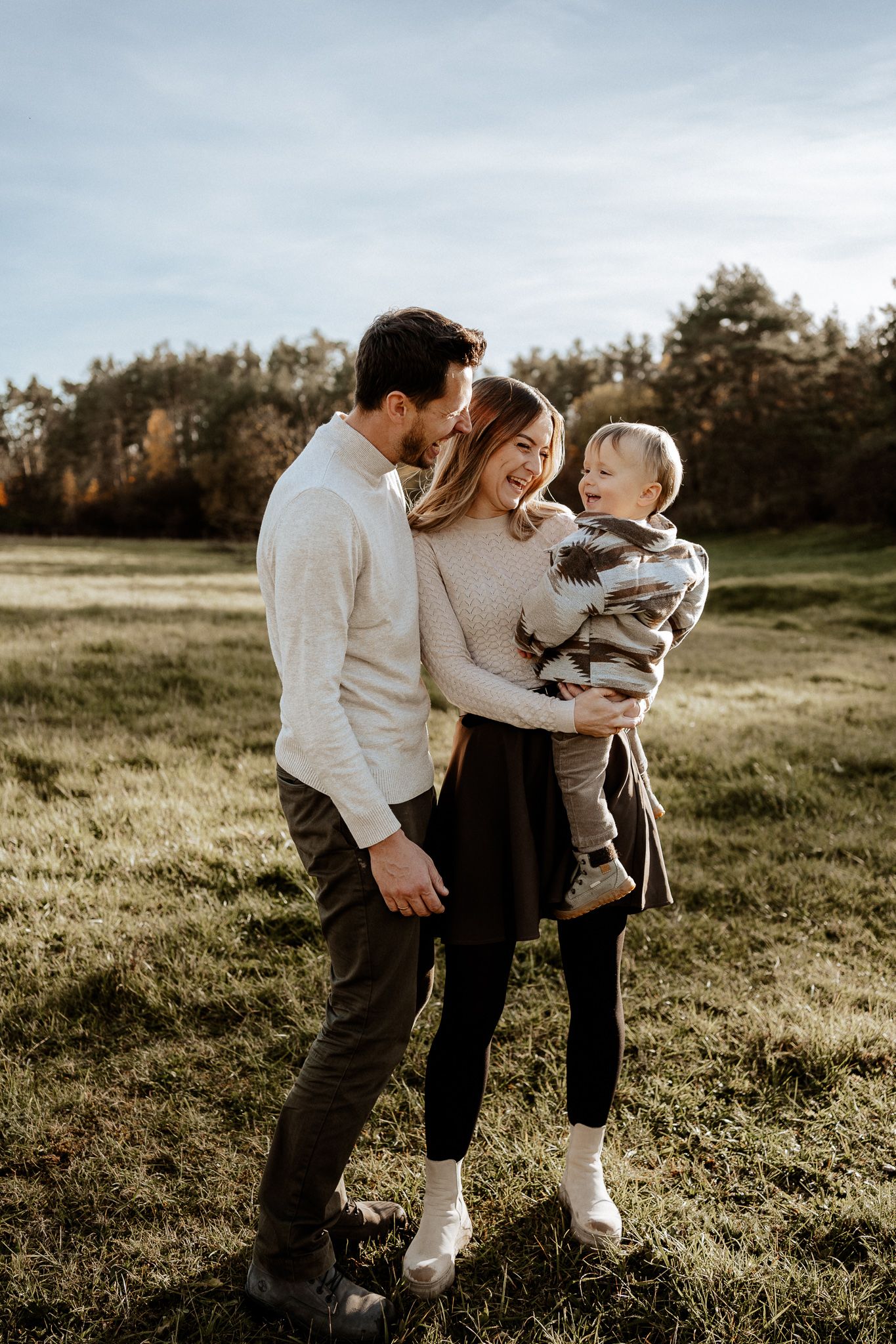 Familie mit Kleinkind lacht gemeinsam draußen auf einer Wiese bei einem natürlichen Fotoshooting.