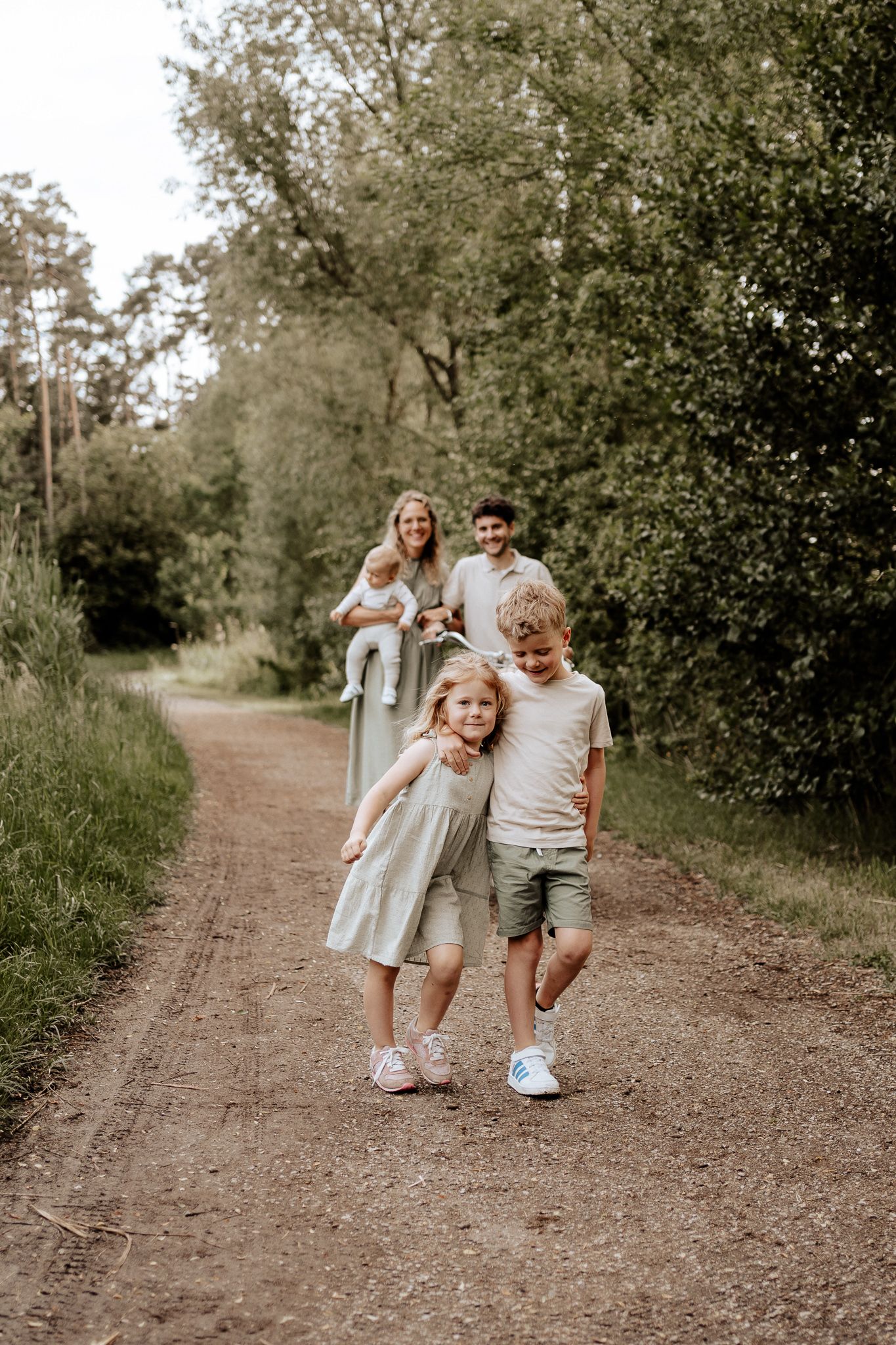 Natürliche Familienfotos beim Spaziergang – Geschwister laufen Hand in Hand auf einem Weg, Eltern im Hintergrund.