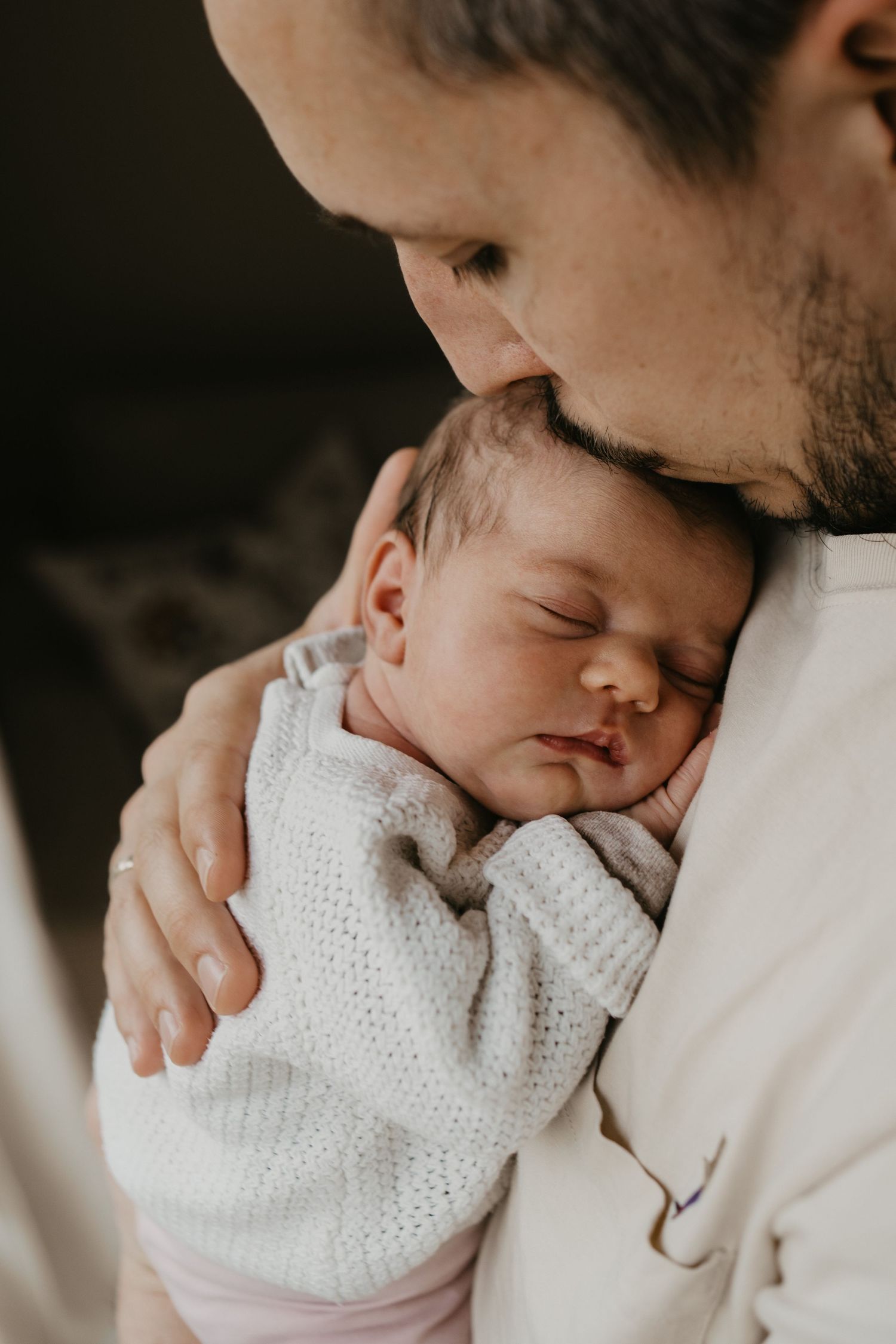 Baby liegt friedlich in den Armen des Vaters. Er küsst das Baby sanft am Kopf während dem Neugeborenen-Shooting in den eigenen vier Wänden.