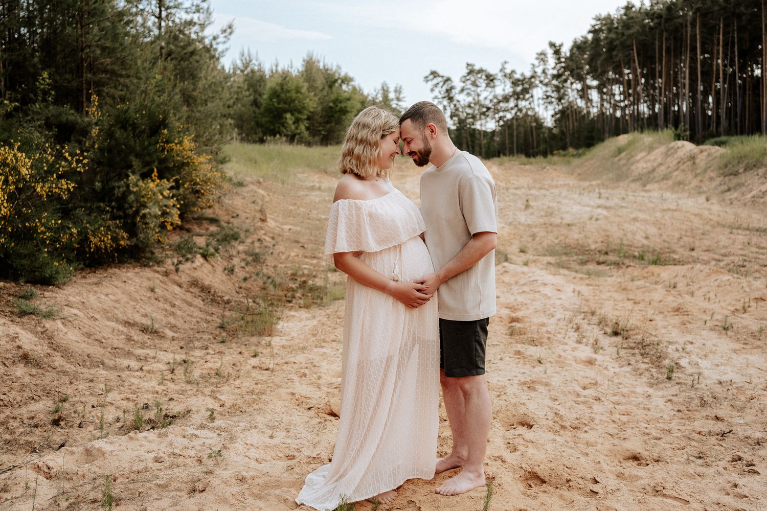 Schwangere Frau in hellem Kleid steht in einer natürlichen Landschaft und hält gemeinsam mit ihrem Partner liebevoll ihren Babybauch.