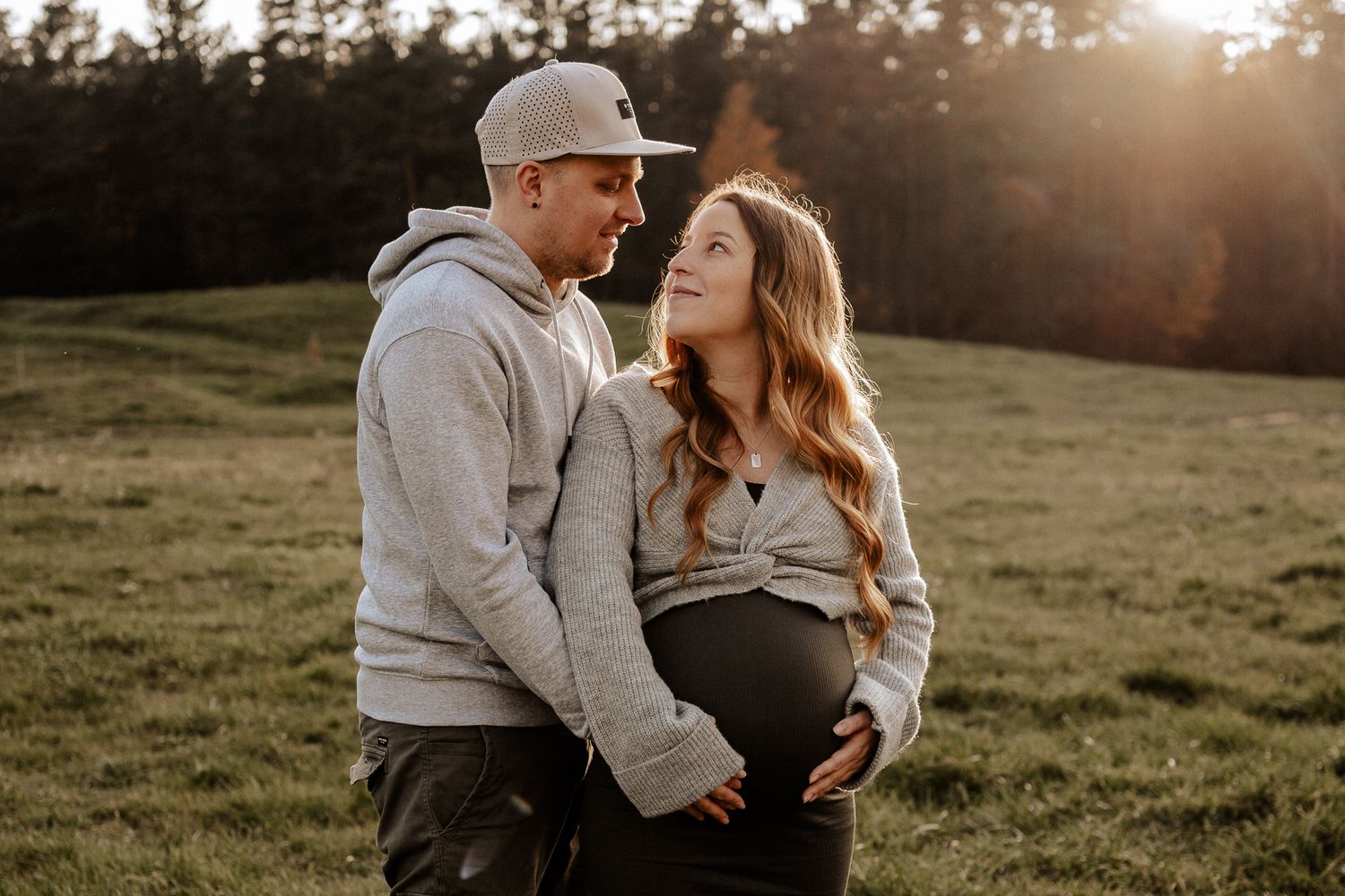Werdende Eltern schauen sich liebevoll an und halten gemeinsam den Babybauch auf einer Wiese.