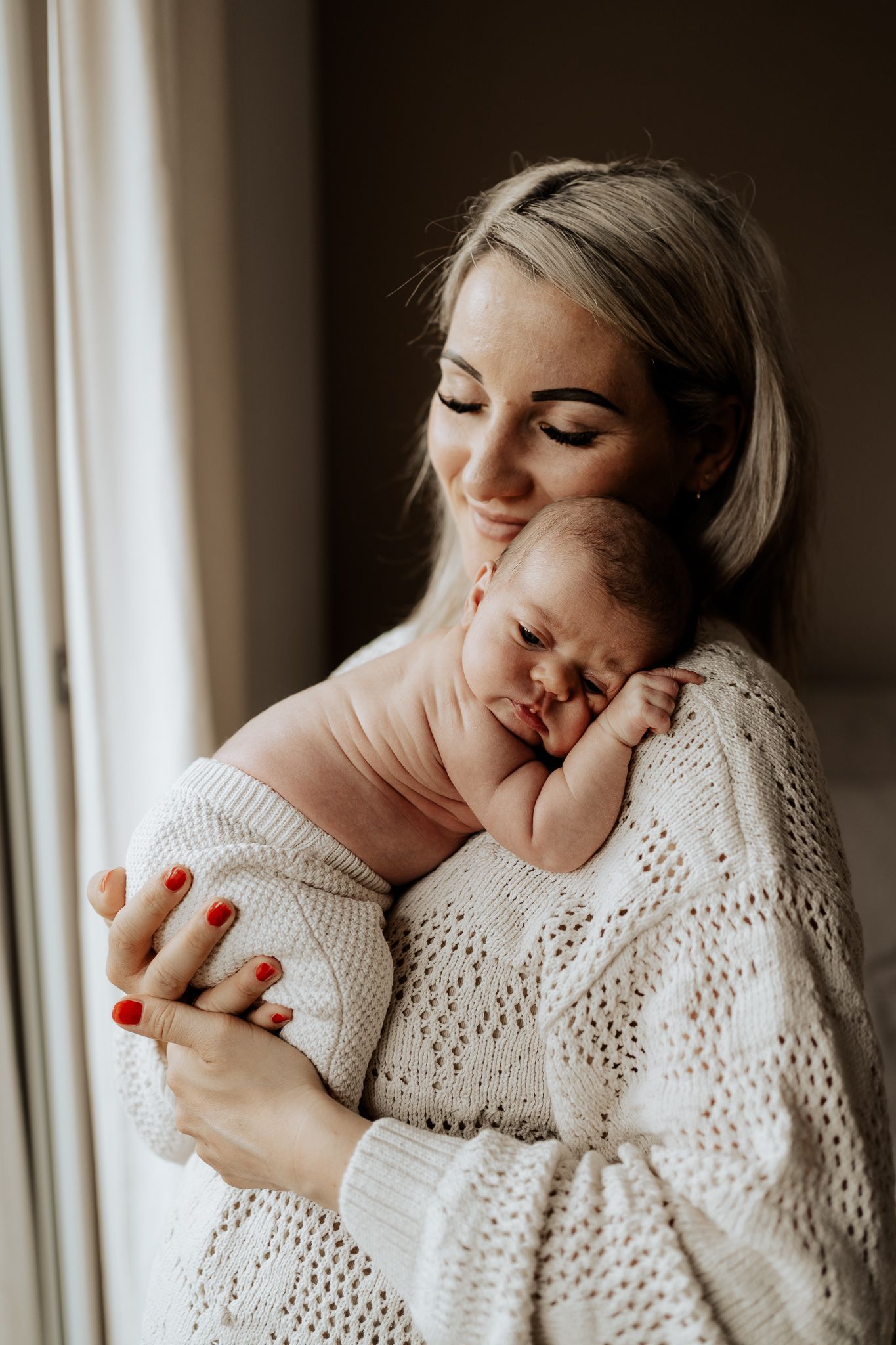 Mutter hält ihr Baby am Fenster im sanften Tageslicht – natürliche Neugeborenenfotografie.