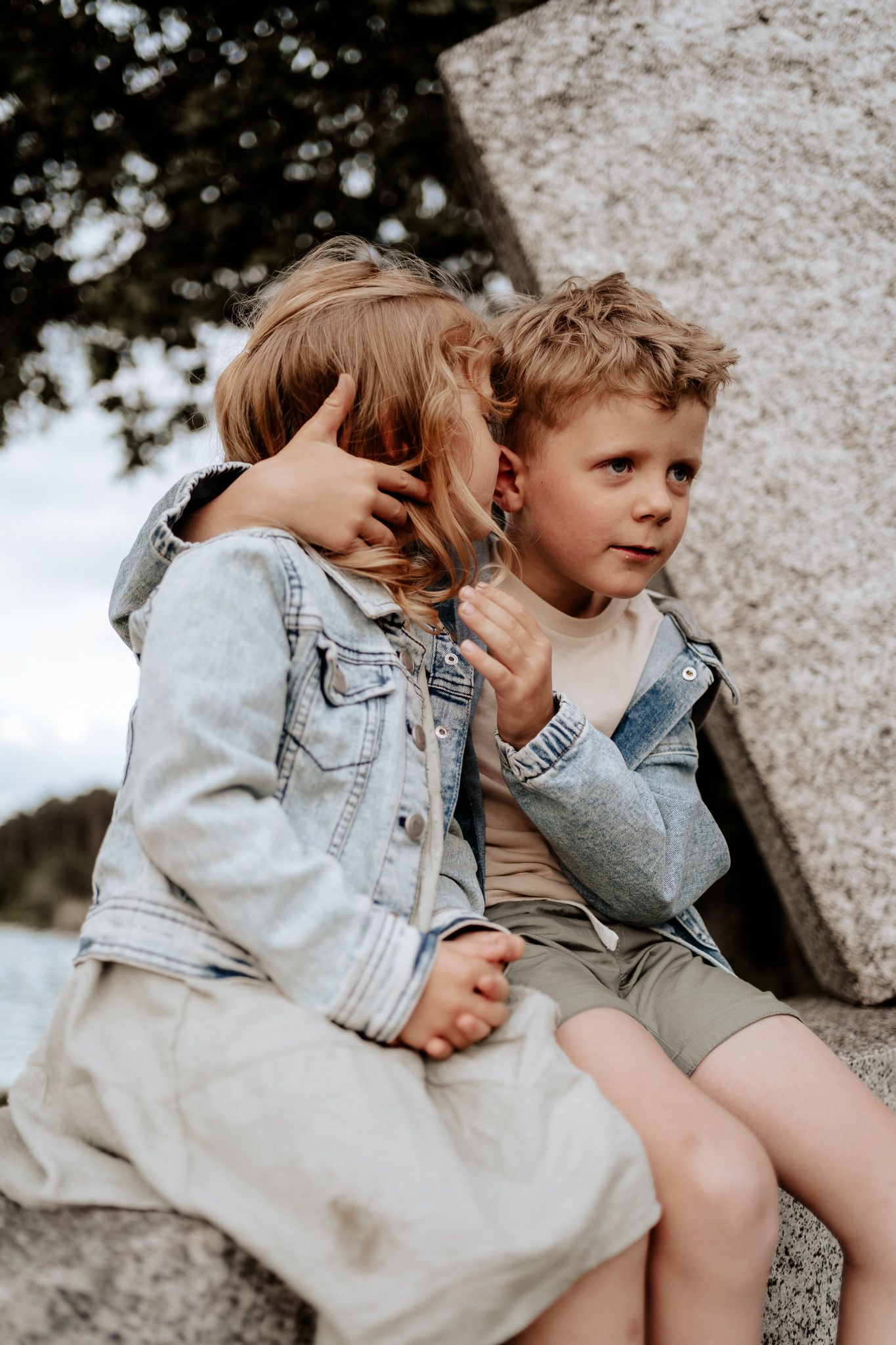 Natürliche Geschwisterfotos – zwei Kinder sitzen eng zusammen und flüstern sich etwas zu.