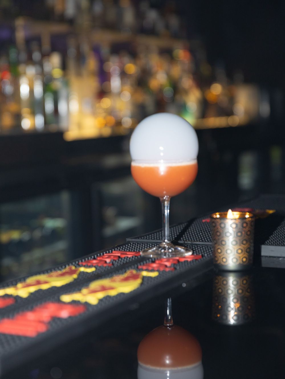 Elegant cocktail with orange liquid and white foam served at a dimly lit bar.