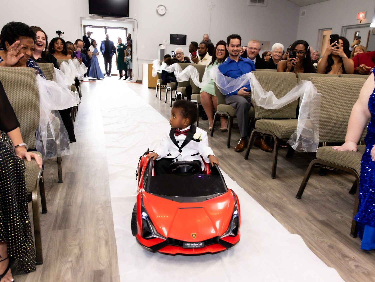 Young ring bearer drives red toy car down aisle at wedding ceremony while guests watch.