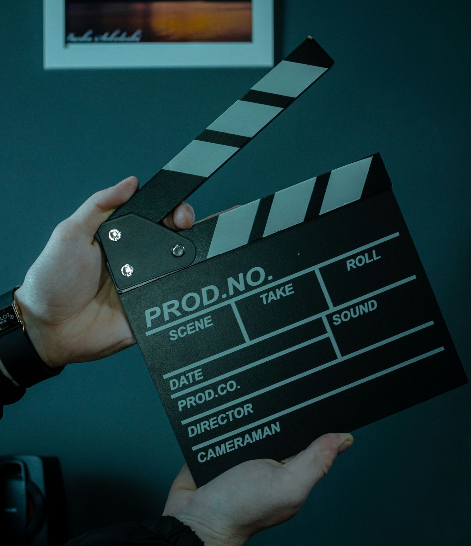 Hands hold a black film production clapperboard in a dimly lit room with teal walls.