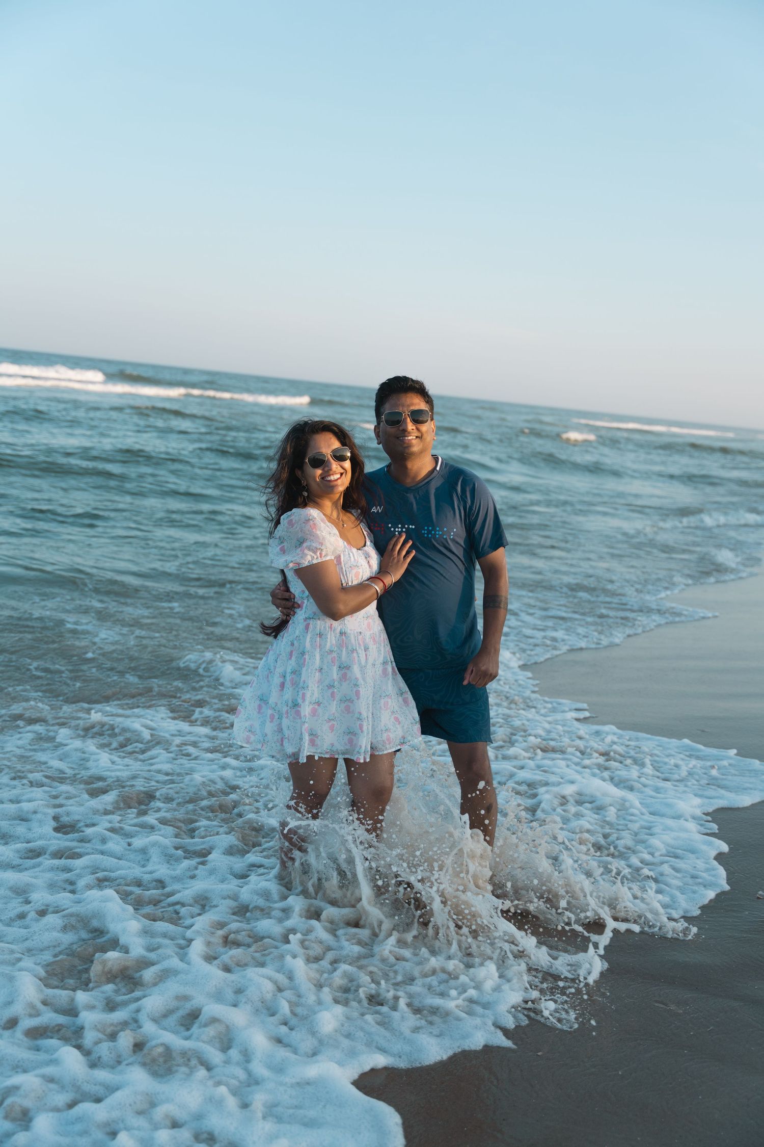 A couple stands smiling together in ocean waves on a sunny beach at sunset.