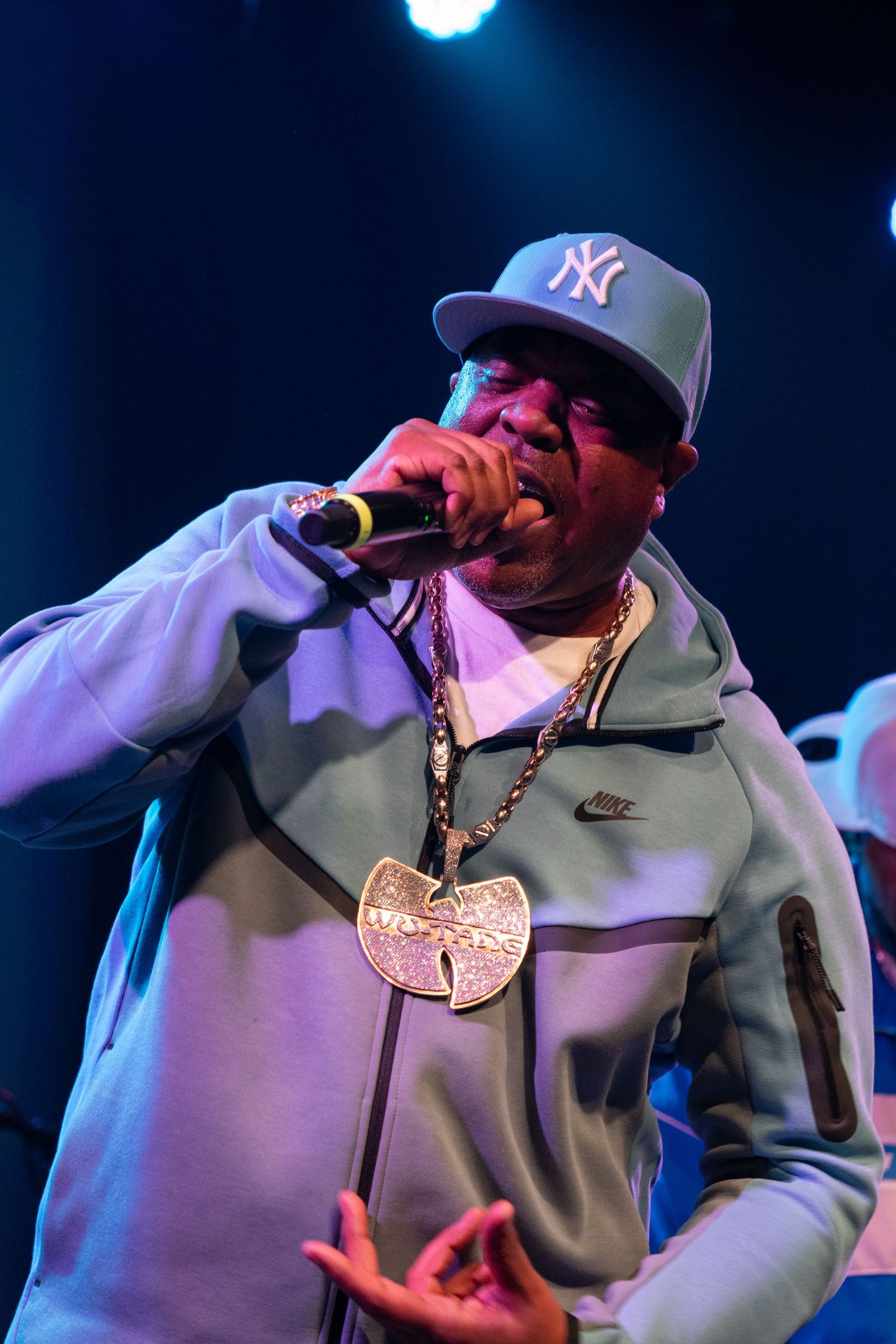 Hip-hop performer in a NY Yankees cap and Wu-Tang chain raps into a microphone on stage.