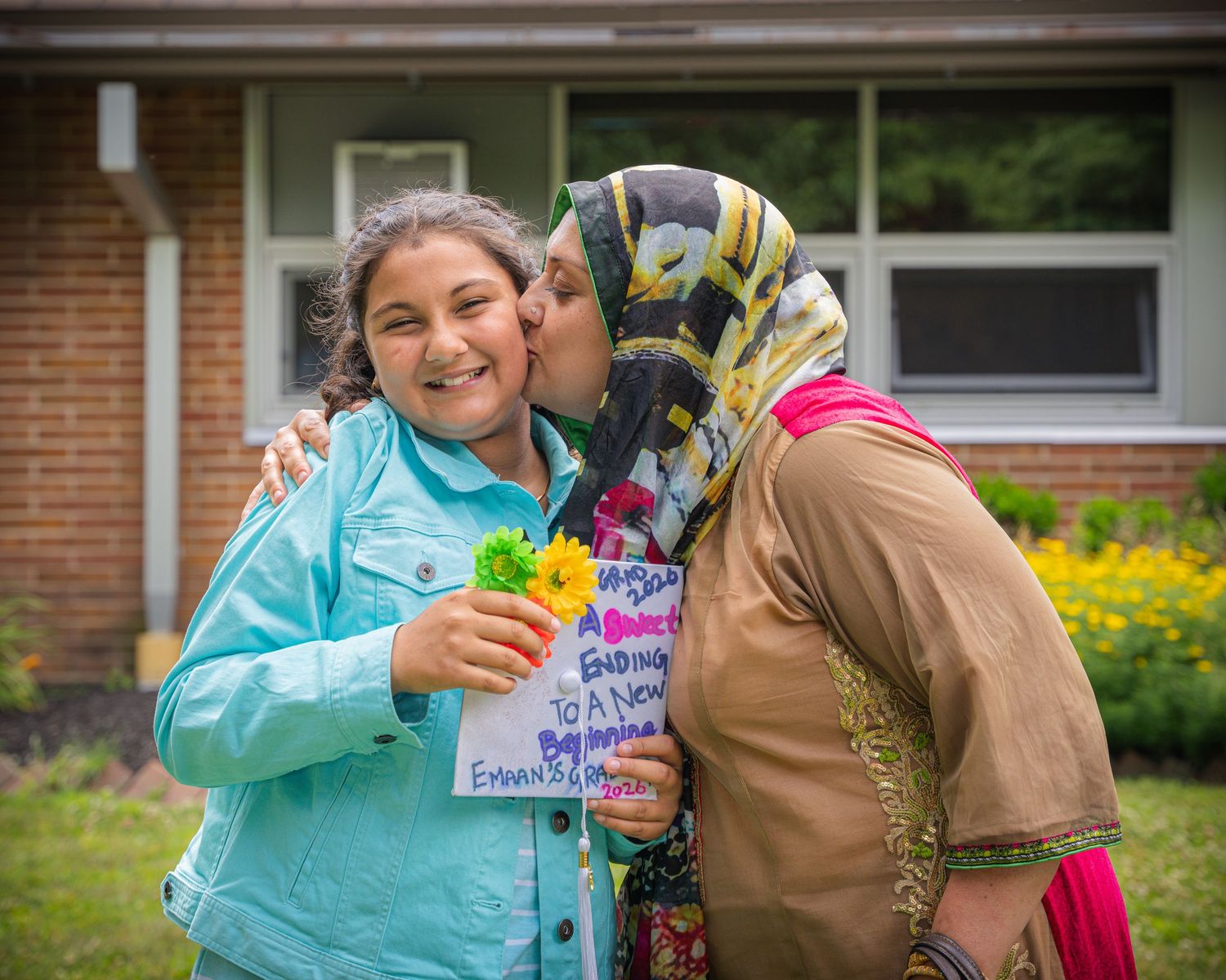 Woman in hijab kisses a smiling girl holding flowers and a congratulations sign outdoors.