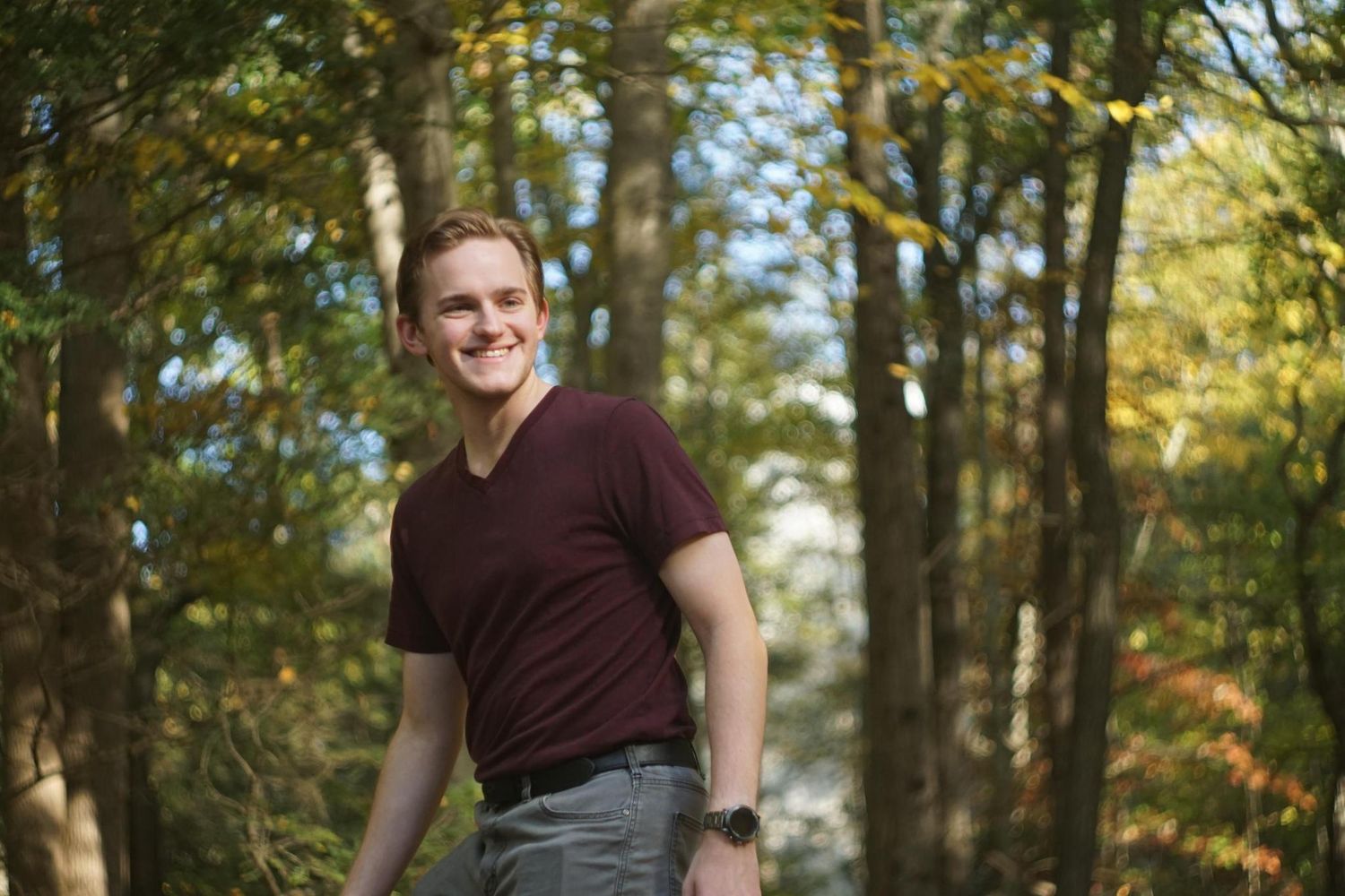 A young man smiles confidently while standing among colorful autumn trees in a forest.