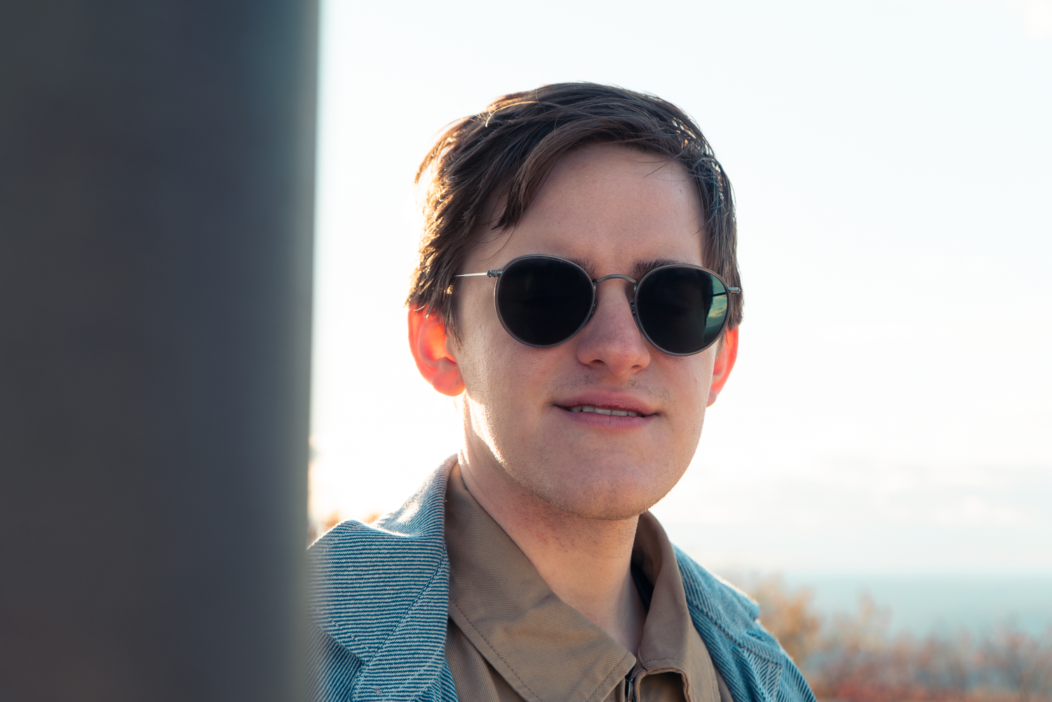 A young person wearing round sunglasses and a light blue blazer smiles outdoors on a sunny day.