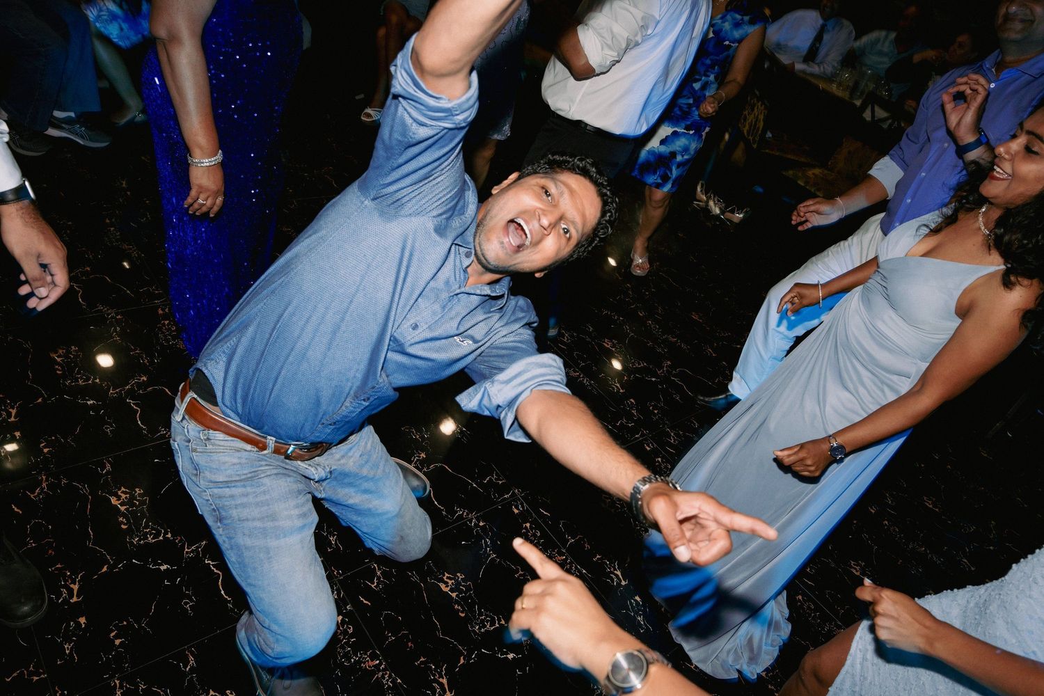 Man in blue shirt dancing energetically at a party with guests in blue outfits on a dark dance floor.