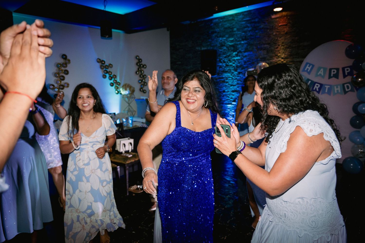 Women dancing joyfully at a birthday party, with one in a blue sequin dress as the focal point.