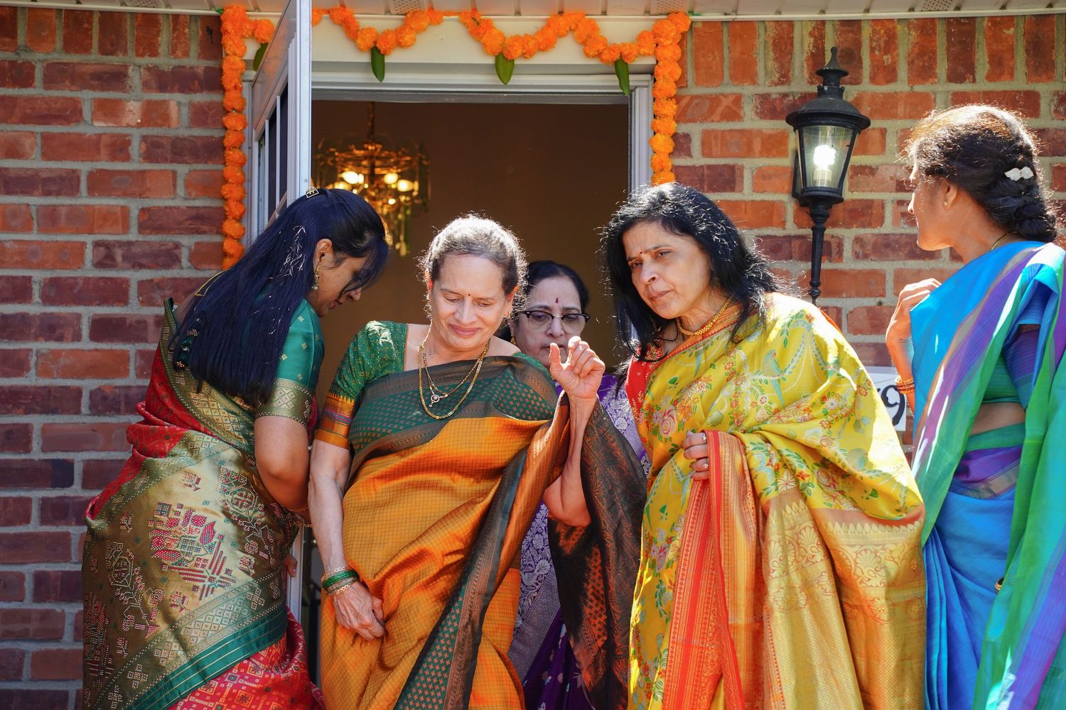 Indian women in colorful silk sarees gather and socialize outside a brick building decorated for a celebration.