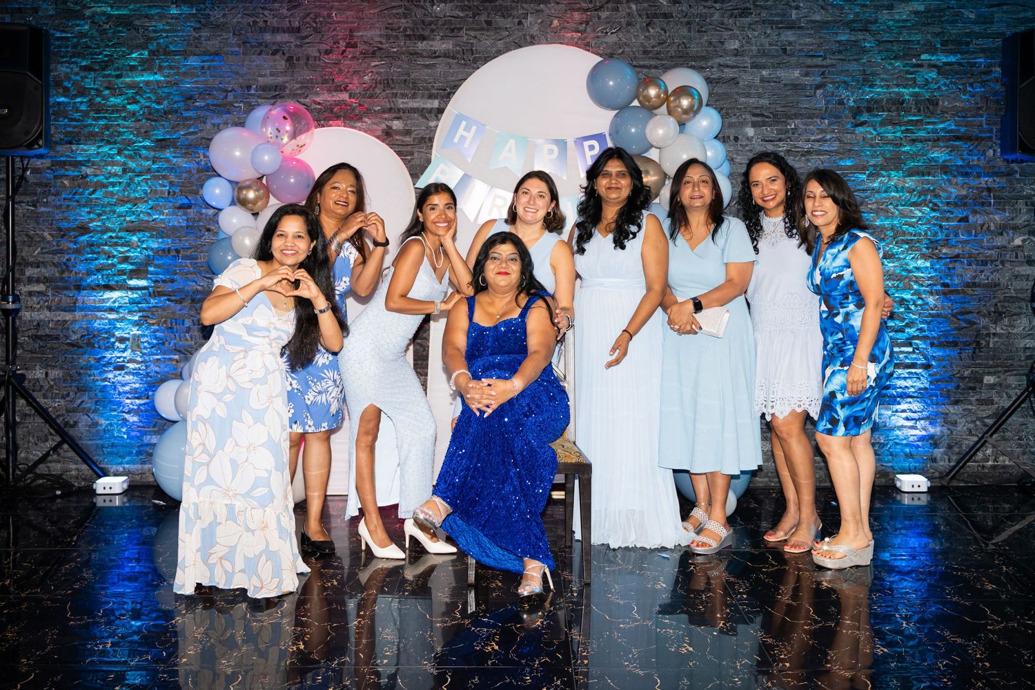 A group of women in blue and white dresses pose together at a festive celebration with balloons.