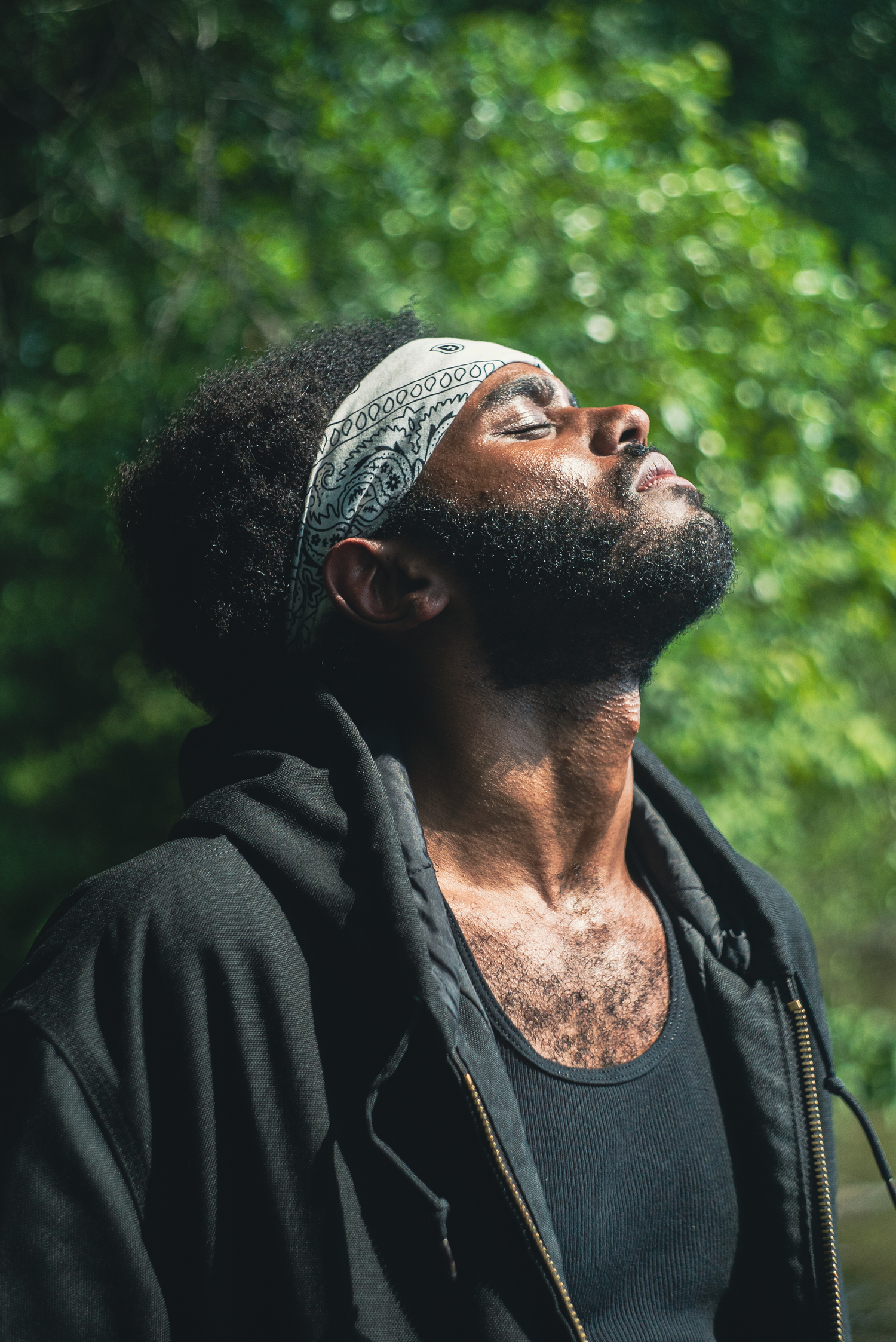 A Black man with a beard and bandana headband tilts his head back with eyes closed outdoors.